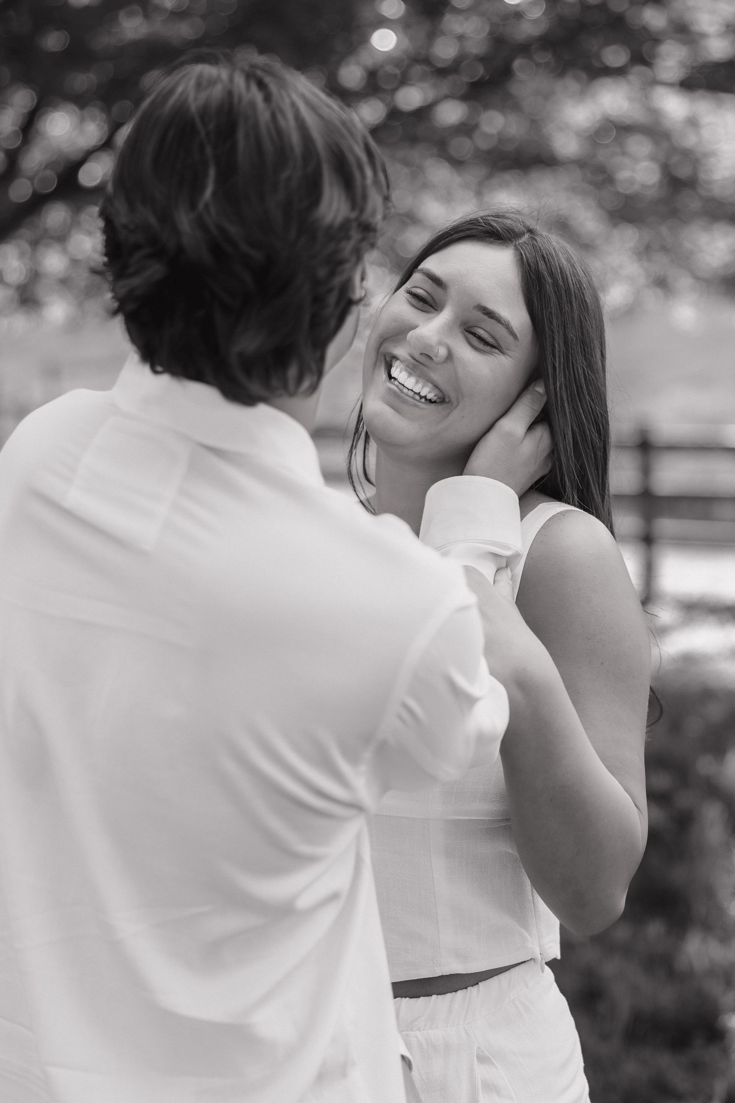 Black and white photo of laughing woman as man tenderly holds her face in a lush outdoor setting.