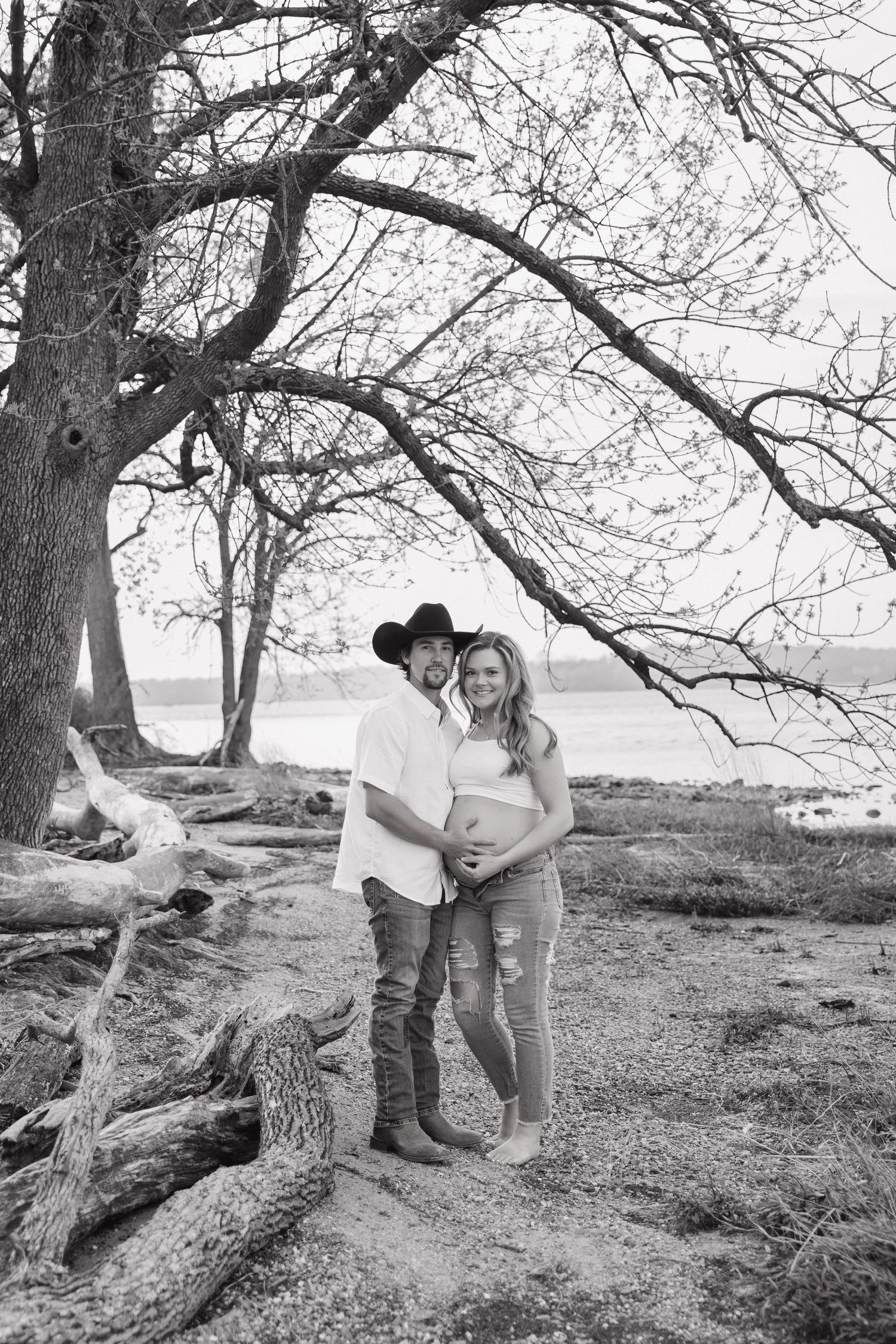 Black and white maternity photo of couple under bare tree branches near a lake, man wearing cowboy hat.