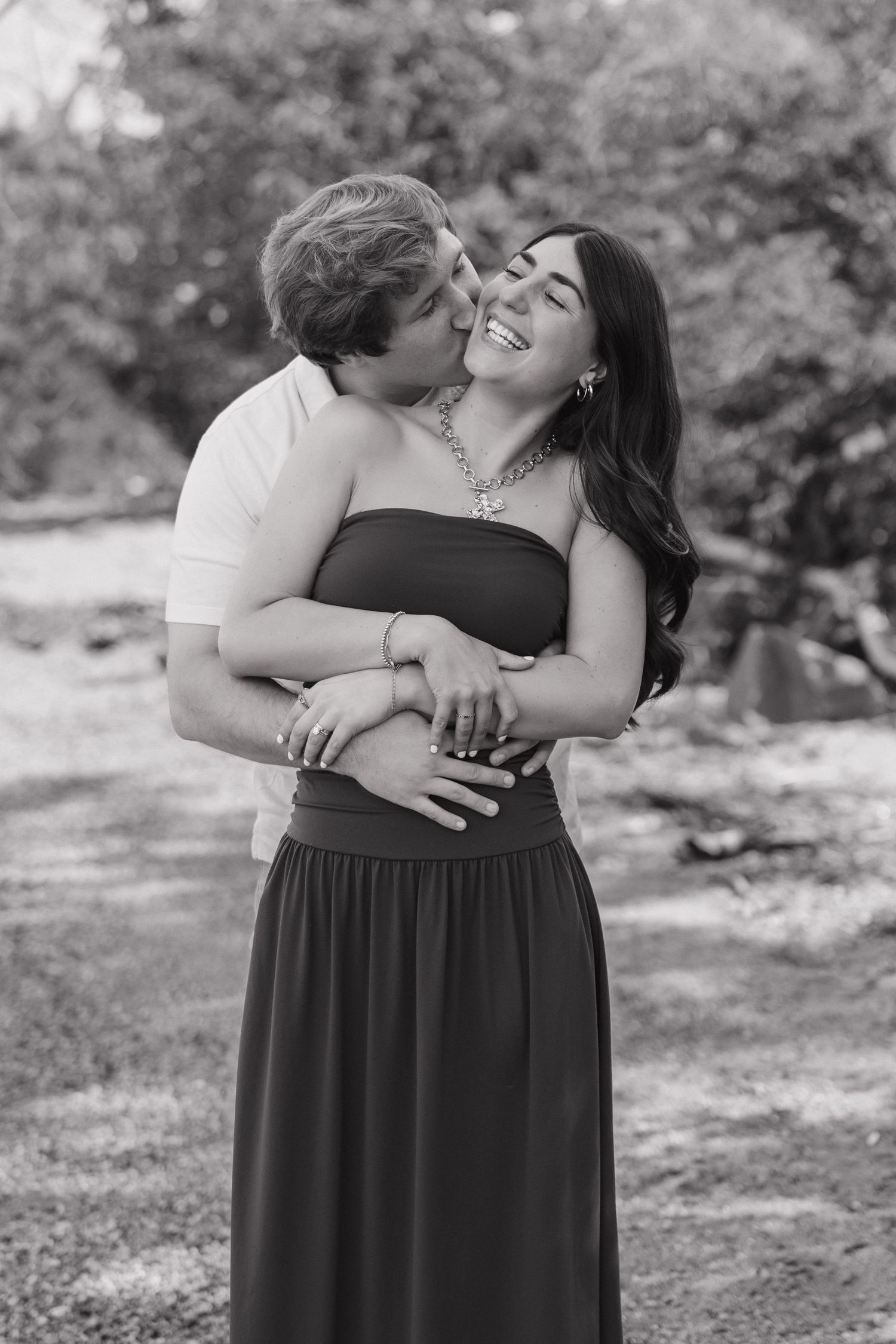 Black and white photo of a couple embracing, man kissing womans cheek as she laughs joyfully outdoors.