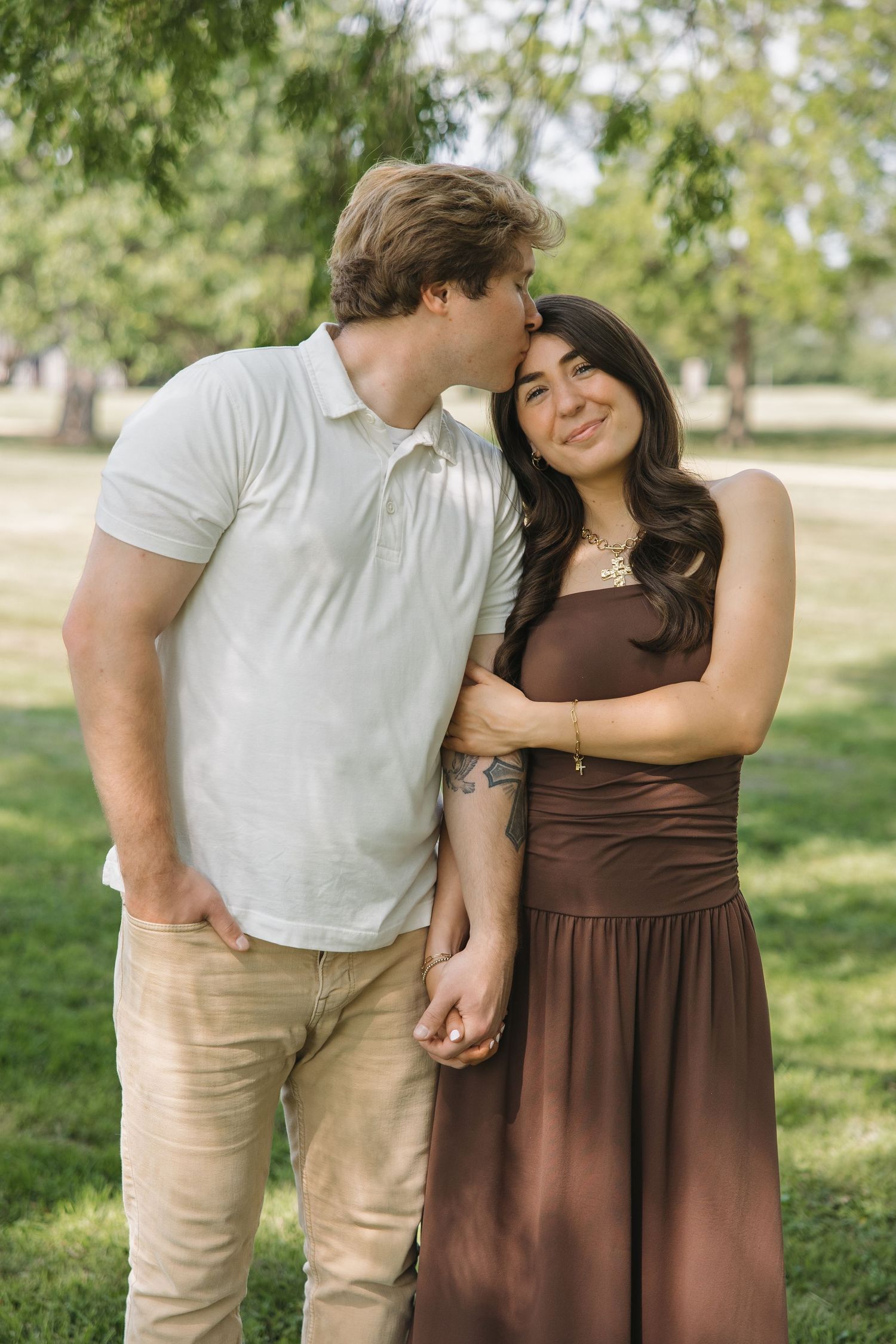 Man kissing womans forehead while she smiles holding hands in a green park.