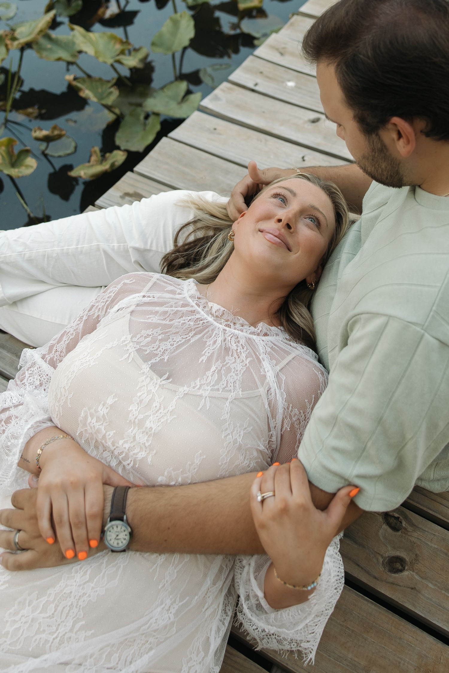 Woman in white lace dress lying on a wooden dock while her partner gently holds her near a lily pond.