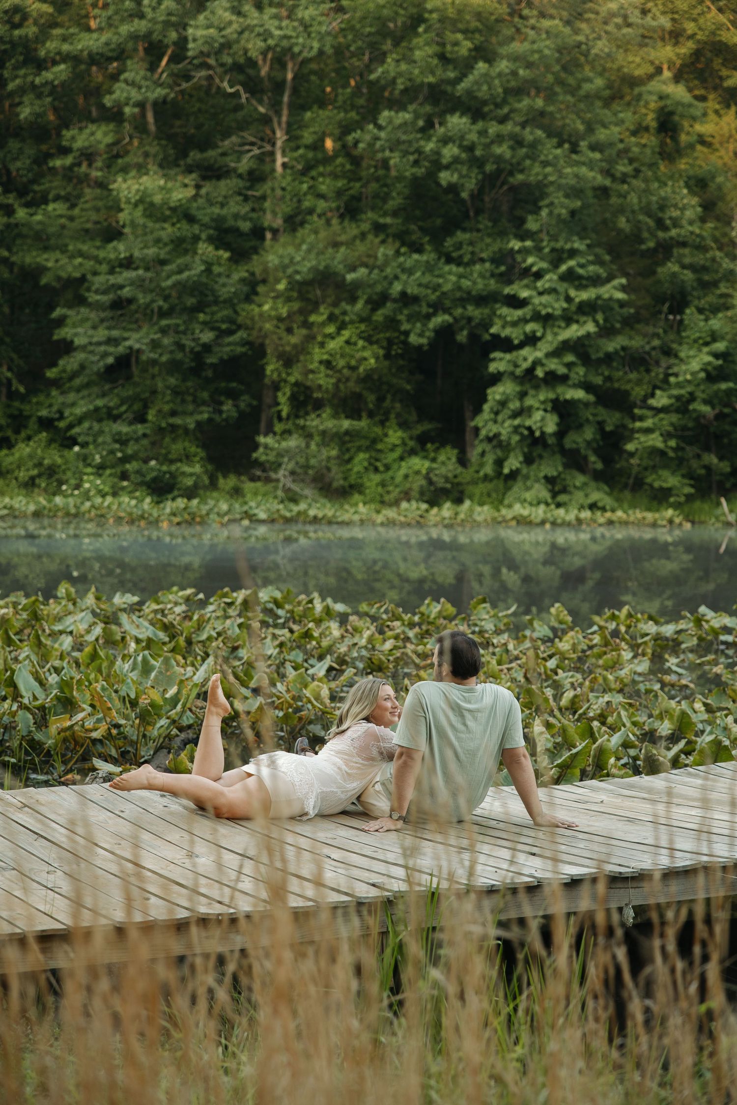 Couple relaxing on wooden dock by serene lake surrounded by green foliage, woman in white dress lying beside man.