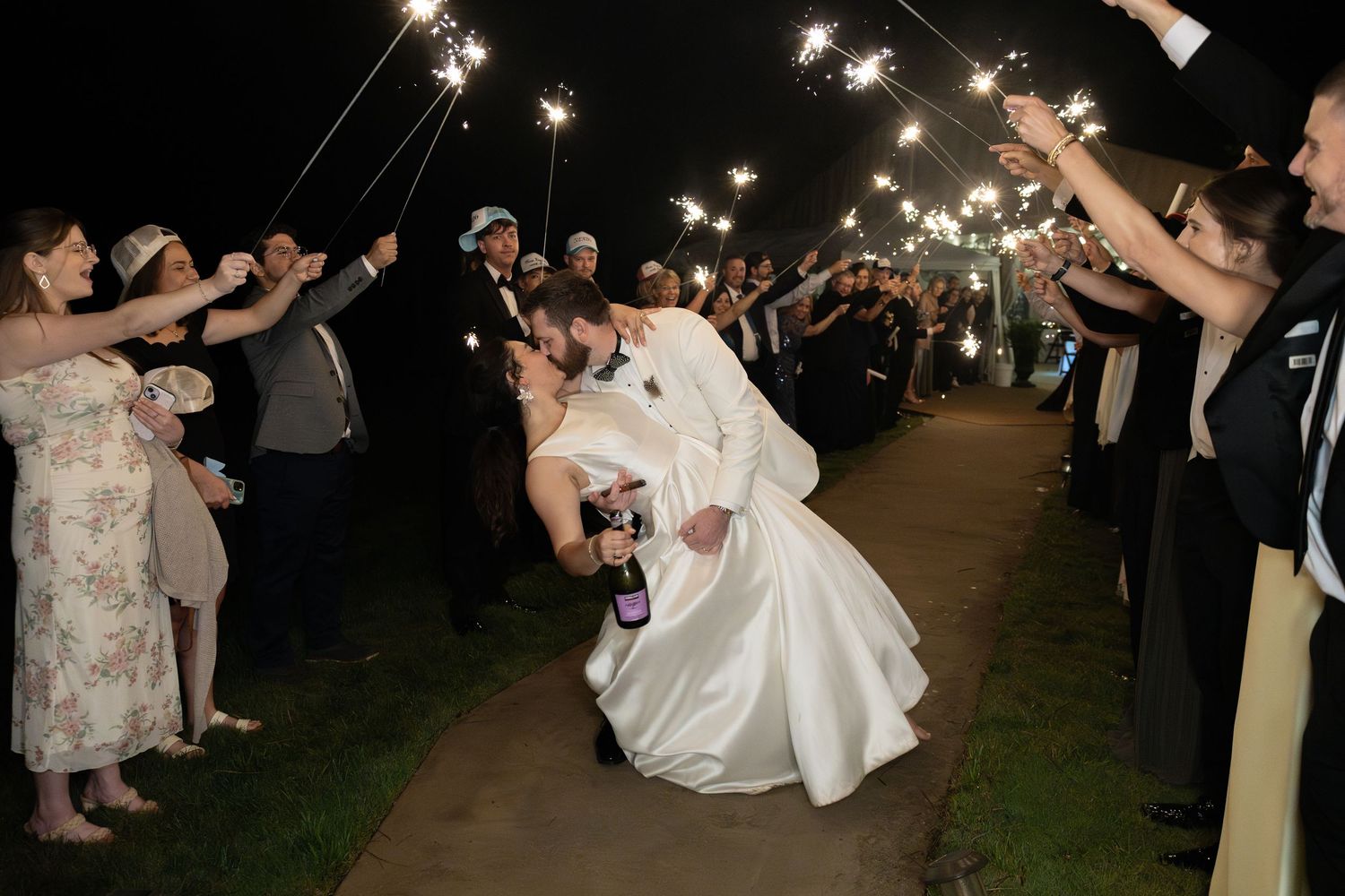 Groom dips bride for a kiss during romantic sparkler exit send-off surrounded by wedding guests holding sparklers at night.