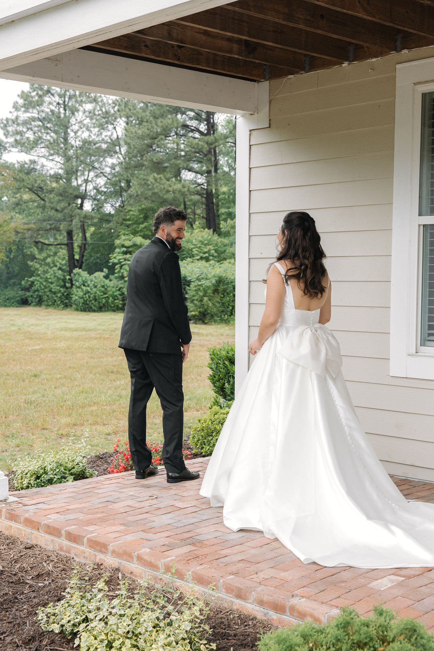 Groom in black tuxedo turns for first look as bride in white ball gown approaches on porch.