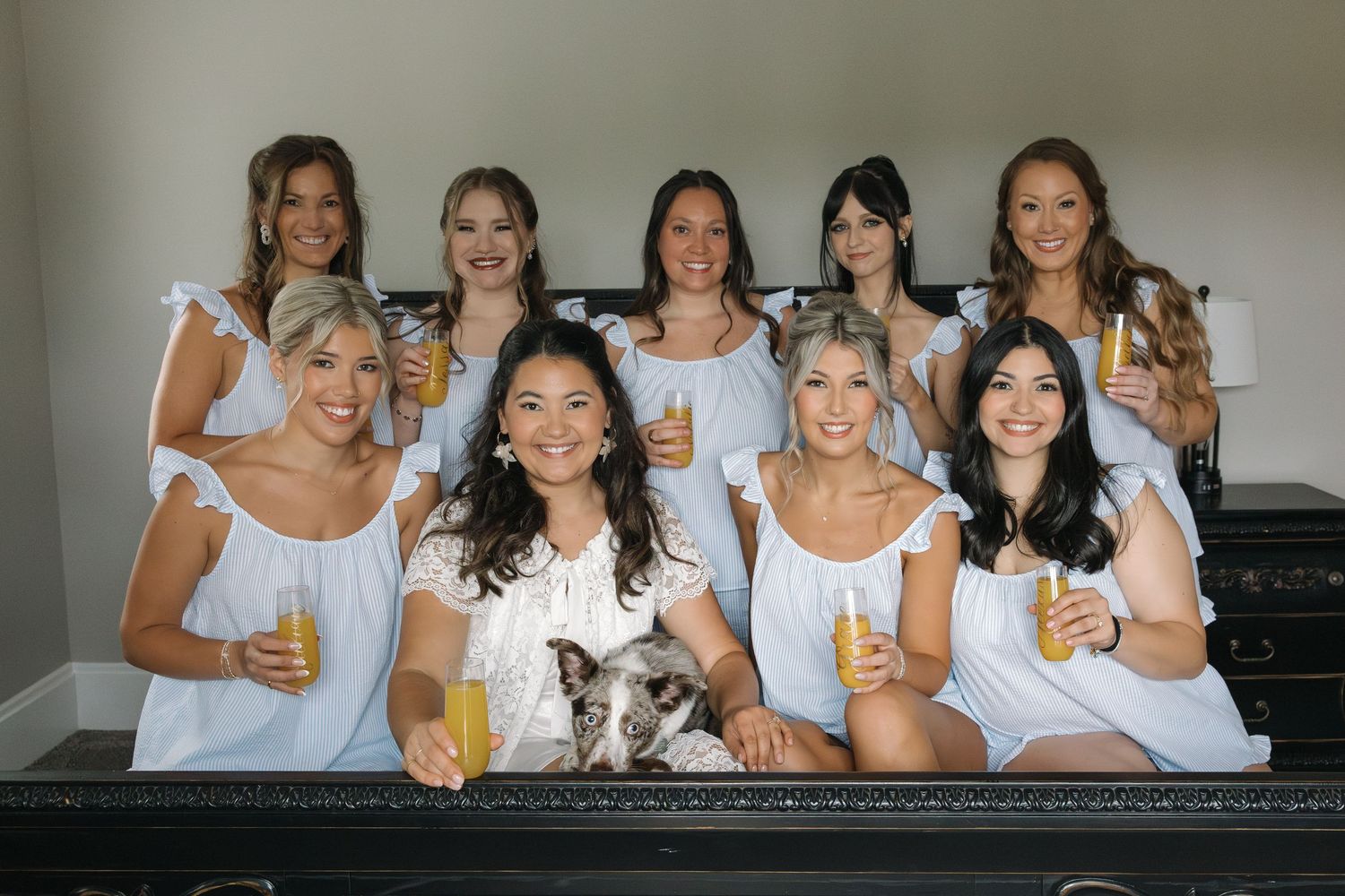 Group of bridesmaids in white dresses toasting with mimosas alongside a small dog.