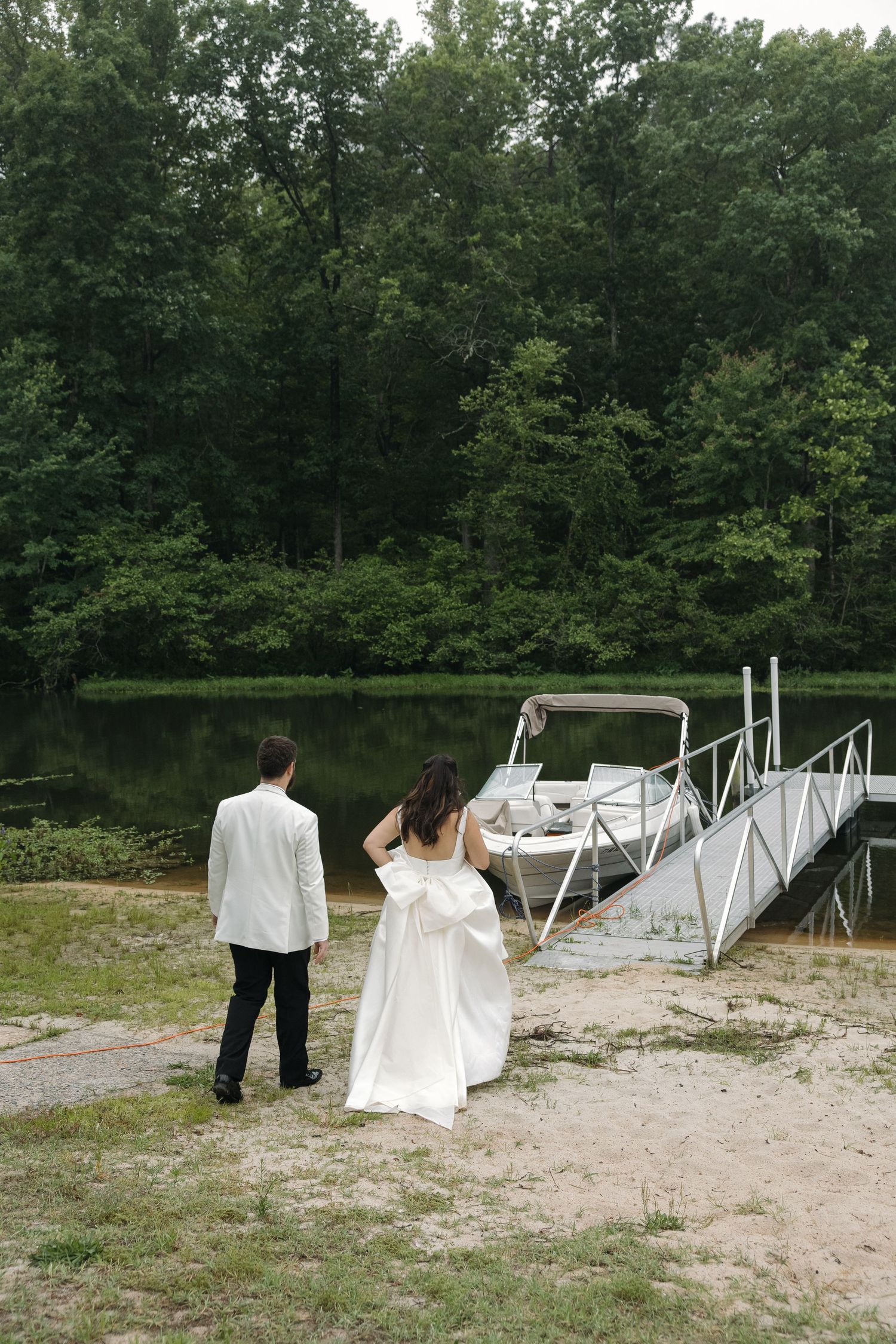 Bride and groom walking toward a white boat dock on a tree-lined lake on their wedding day.