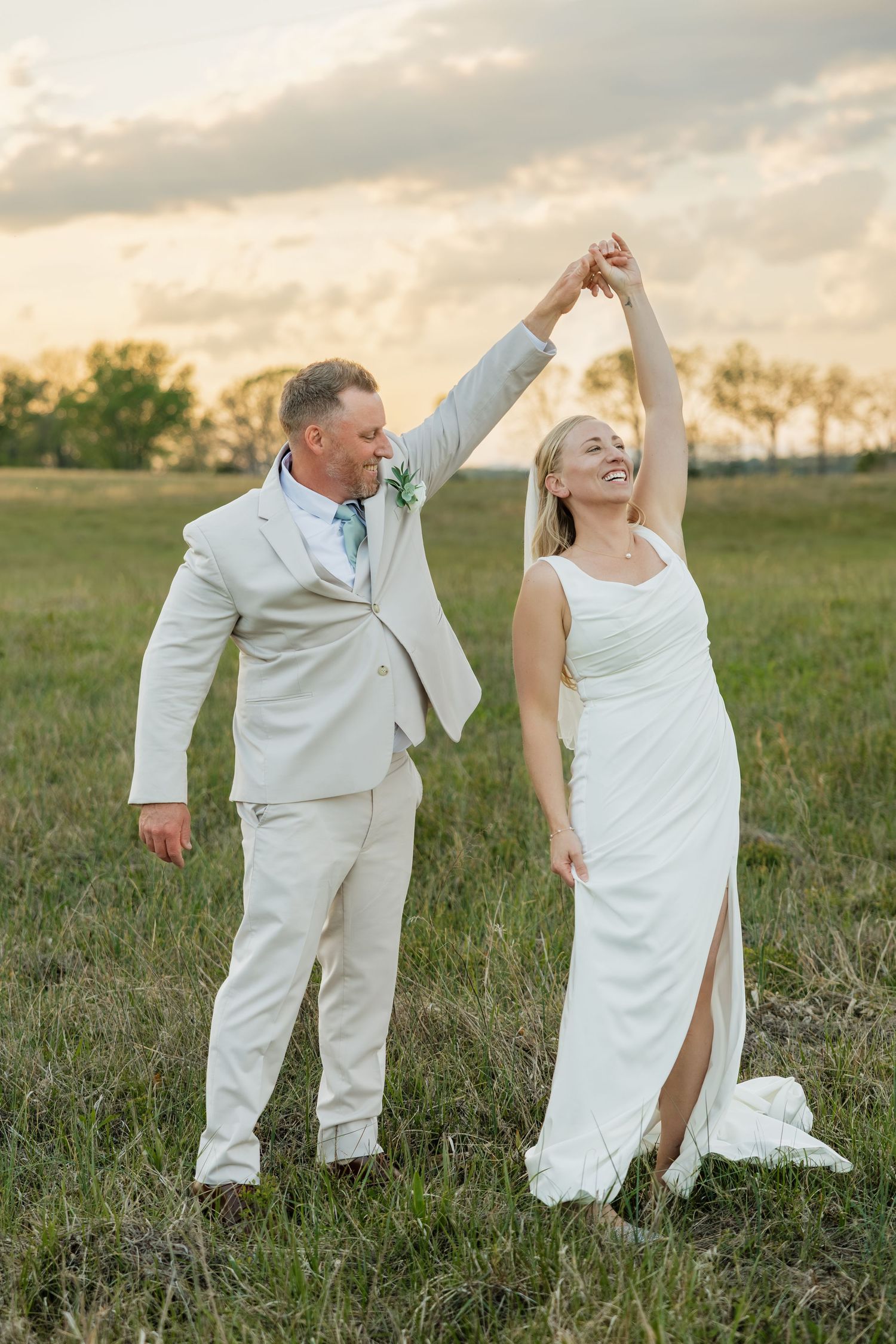 Groom twirls smiling bride in a white gown in an open field at sunset.