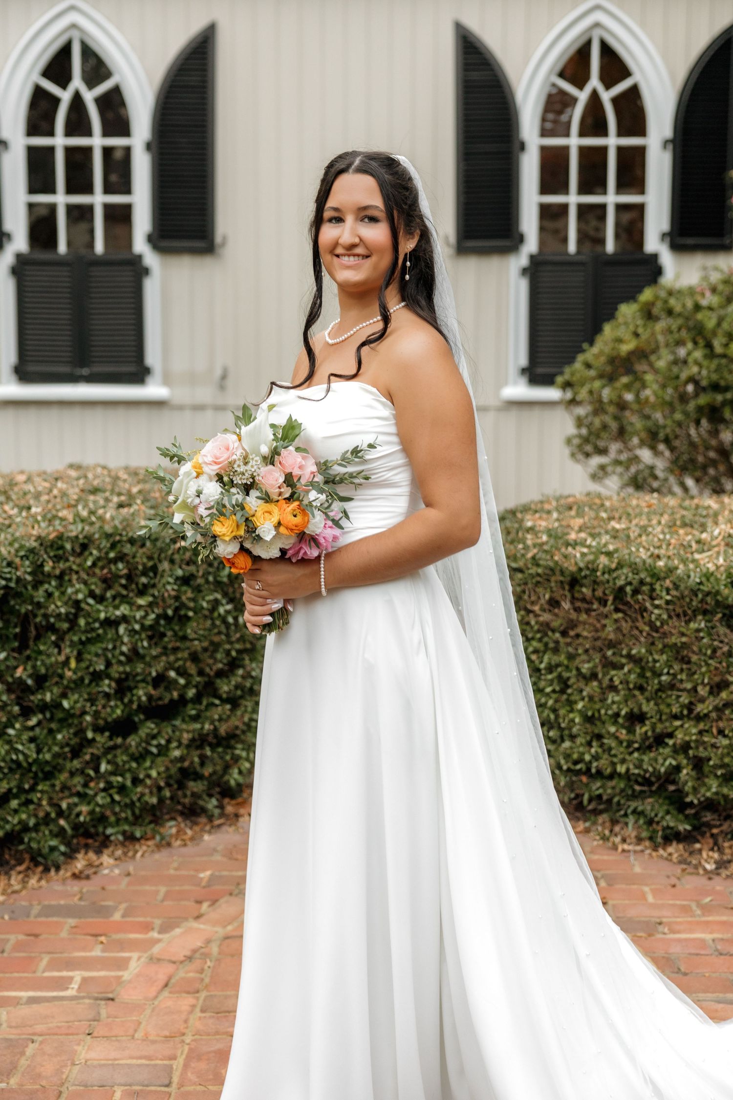 Smiling bride in a strapless white gown with veil holding a colorful bouquet outside a white chapel.