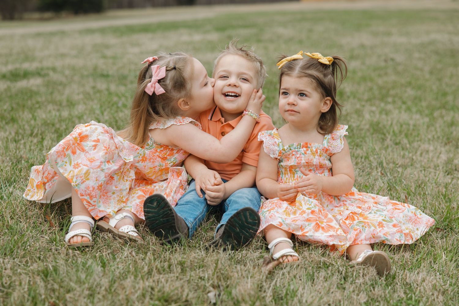 Three young siblings sit on grass, two girls in floral dresses kiss and hug their smiling brother in an orange shirt.