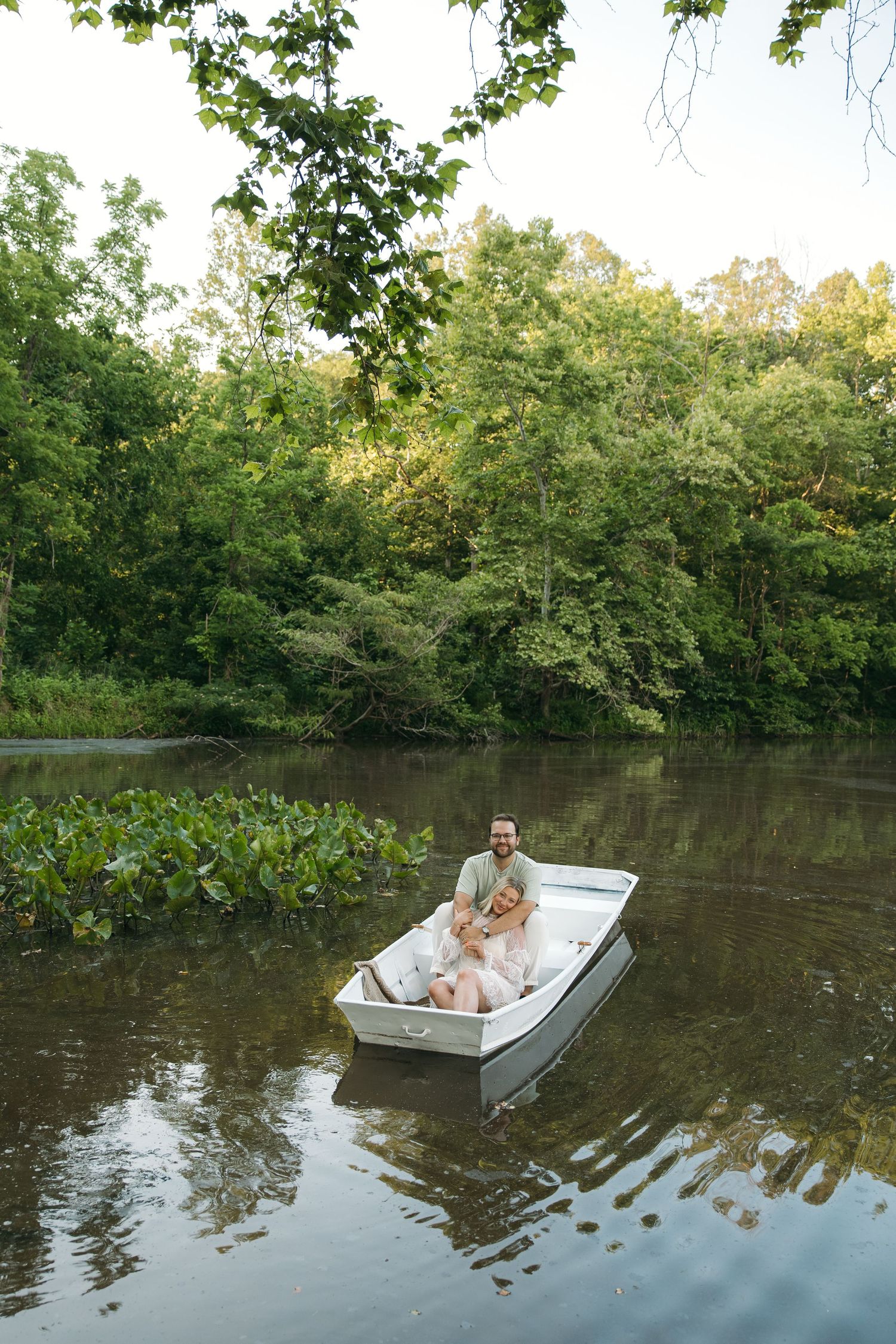 A romantic couple relaxes together in a small white rowboat surrounded by lush green trees on a river.