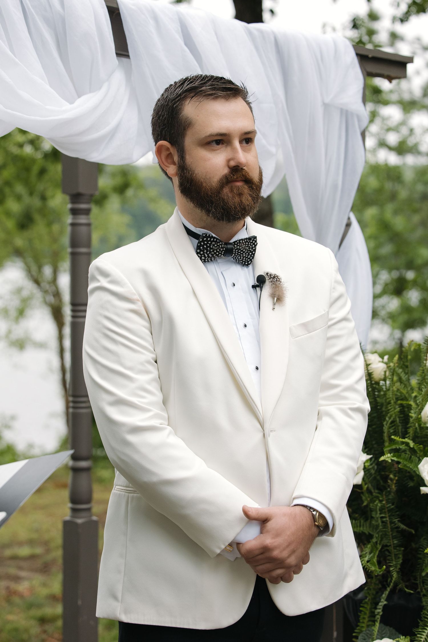 Bearded groom in white tuxedo jacket and black bow tie standing at outdoor wedding altar.