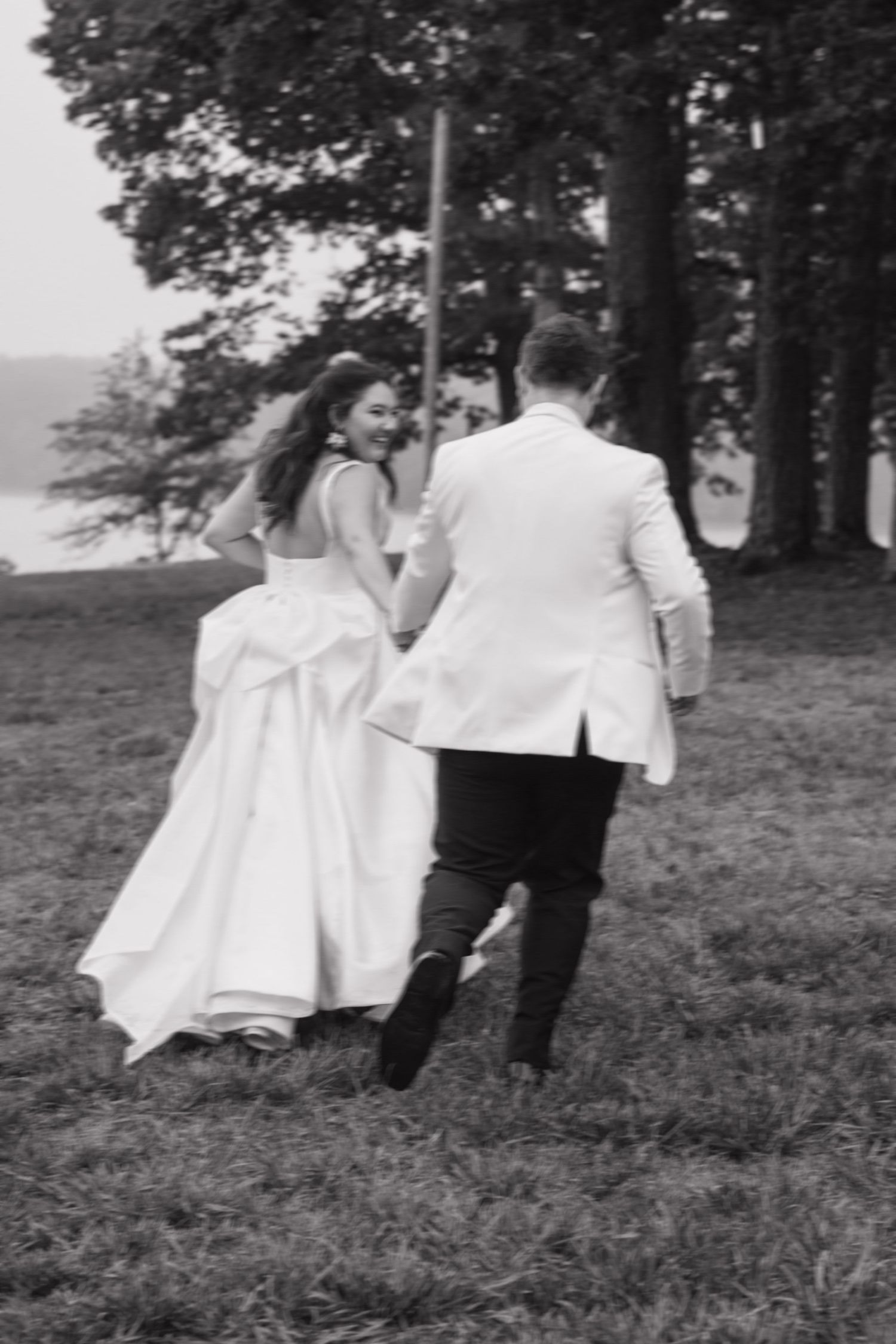 Black and white photo of bride and groom running hand in hand across grassy field.
