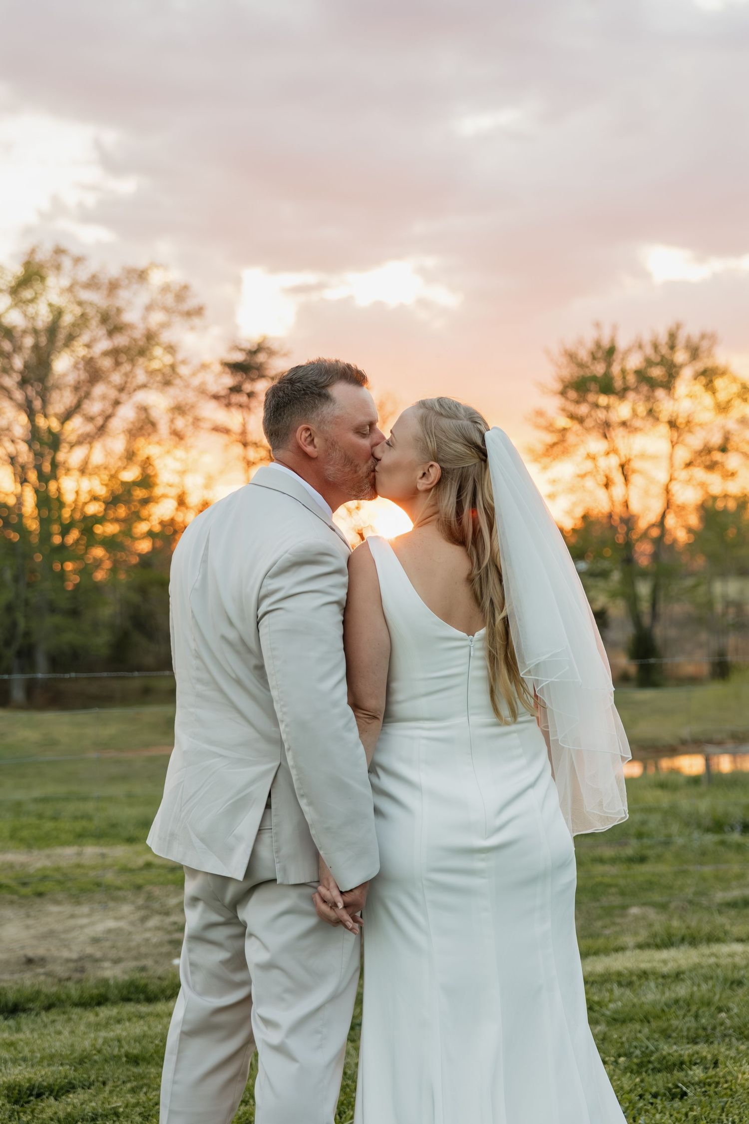 Bride and groom sharing a romantic kiss at golden sunset with soft pink clouds behind them.