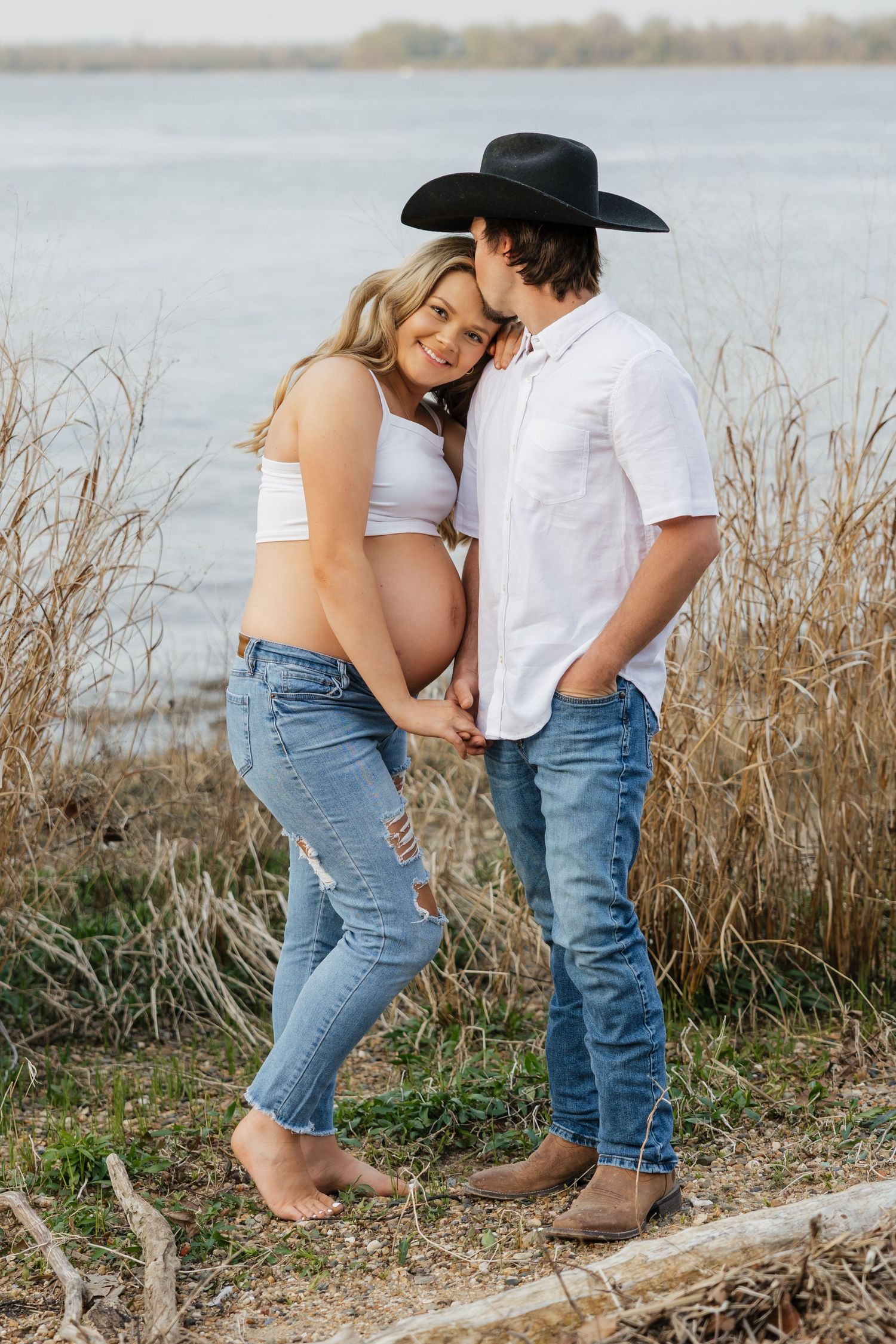 Expectant couple shares tender moment by lakeside reeds, man in black cowboy hat kissing pregnant woman's temple.