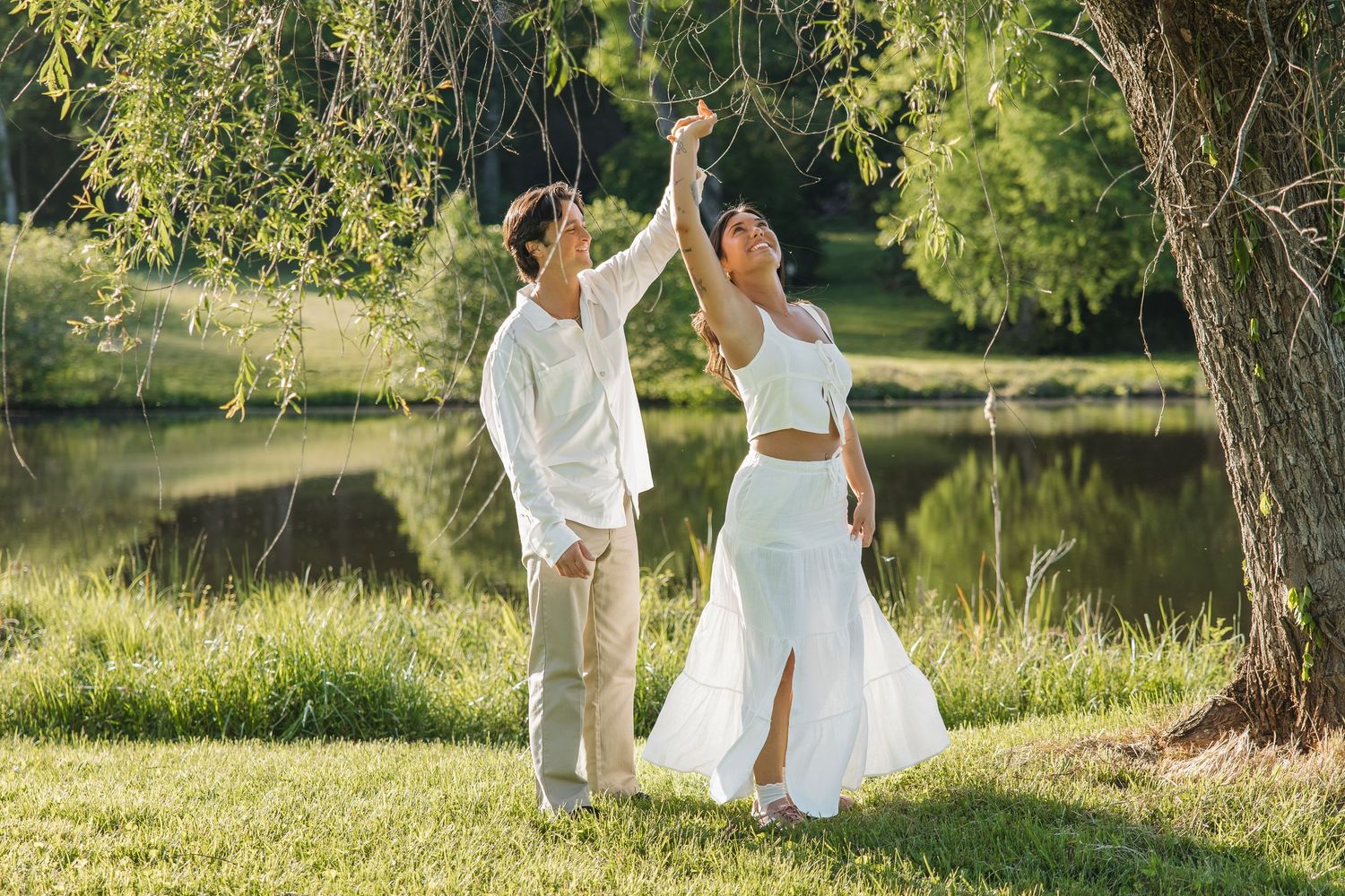 Couple in matching white outfits dancing and holding hands under a willow tree beside a tranquil pond.