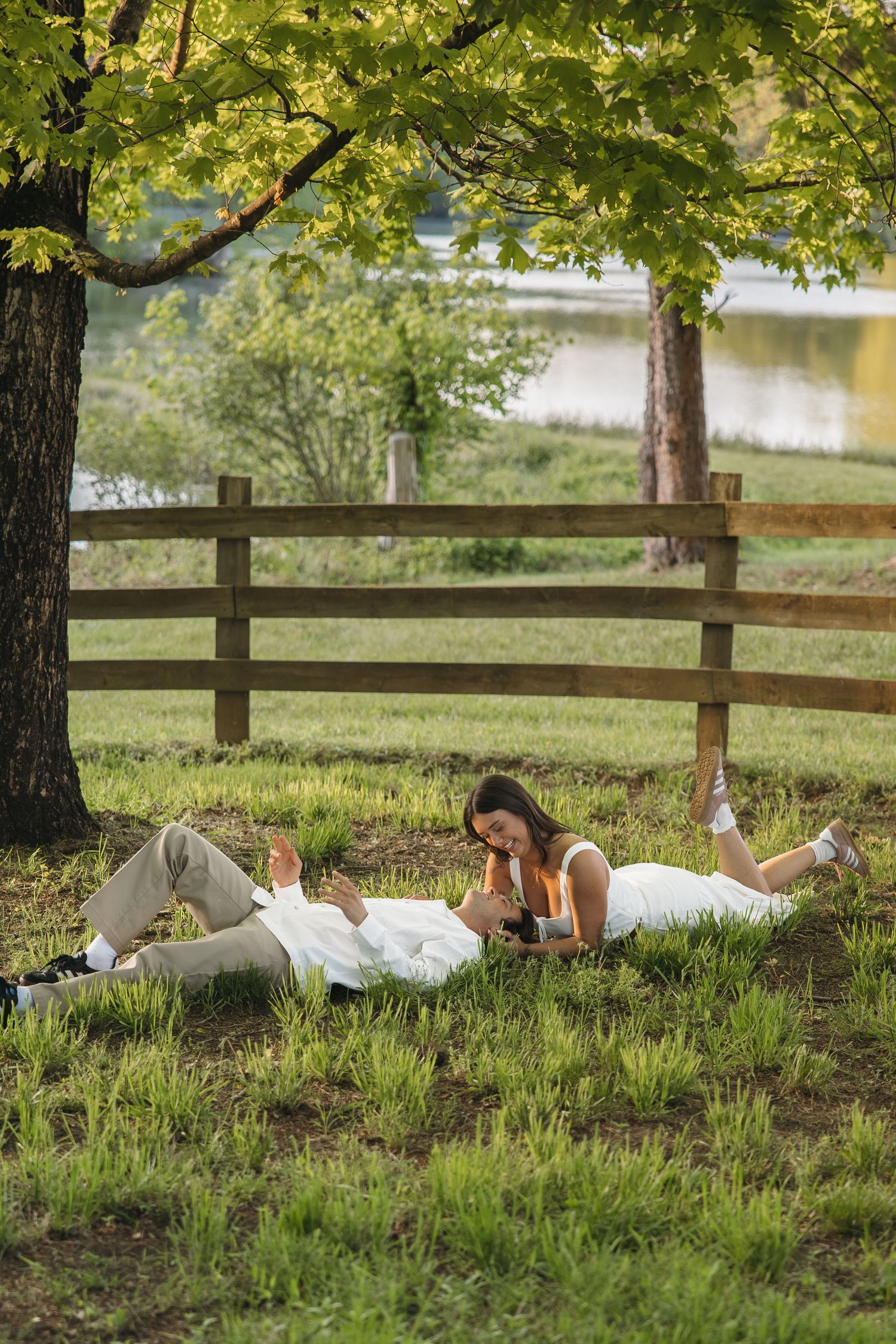 Couple lying on grass under a maple tree near a wooden fence and pond in a lush green park.