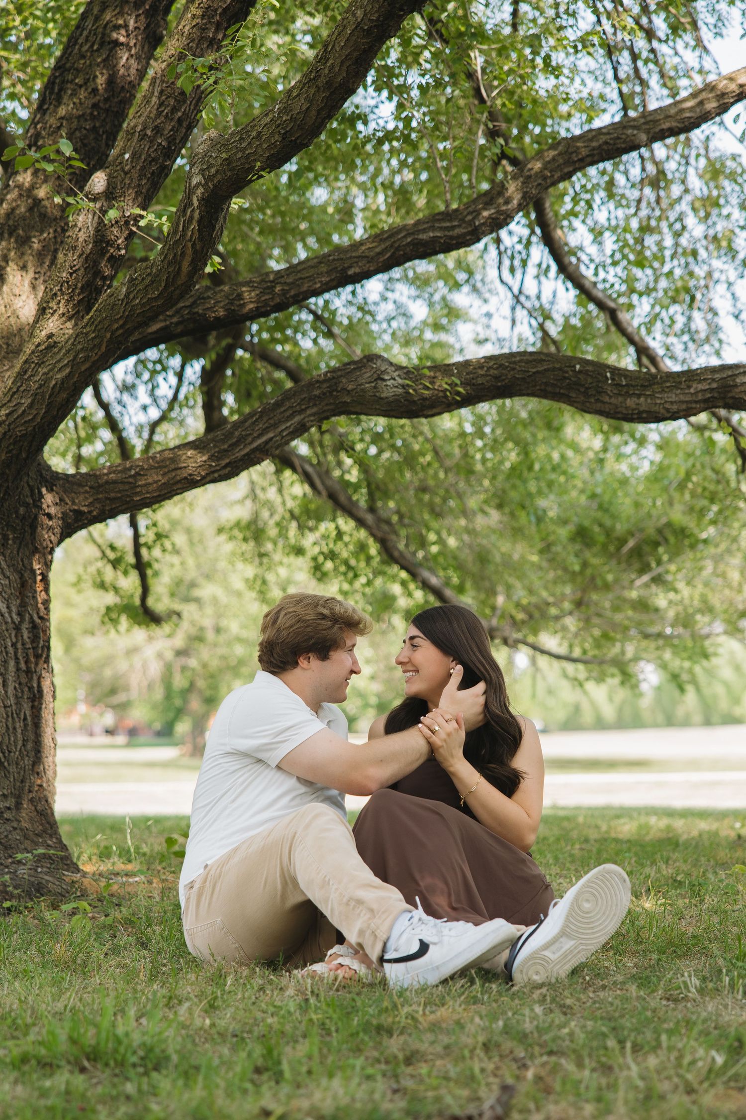 A joyful couple sits under a large tree on green grass, holding hands and smiling at each other.