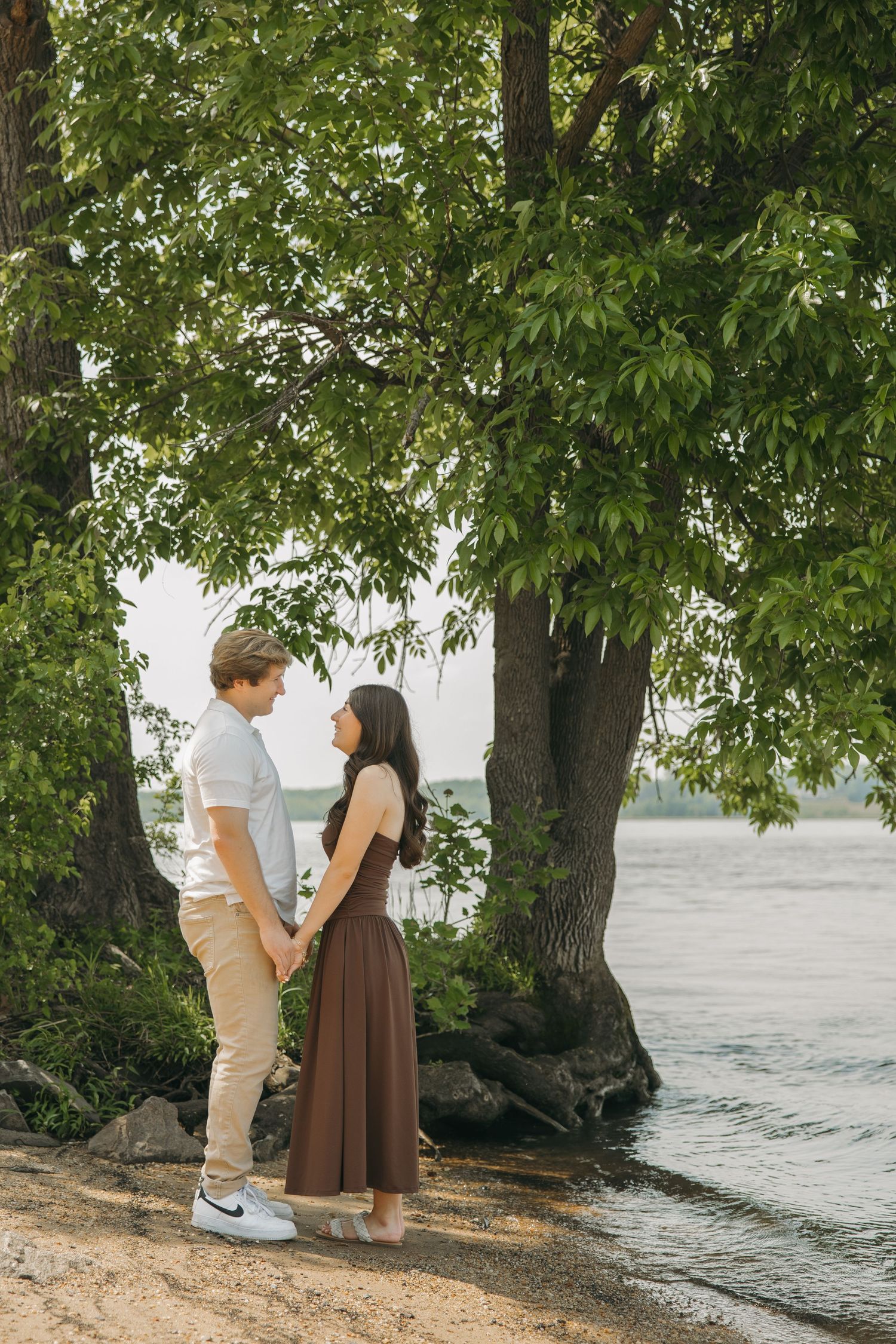 Romantic couple holding hands facing each other under a large tree by a lake.
