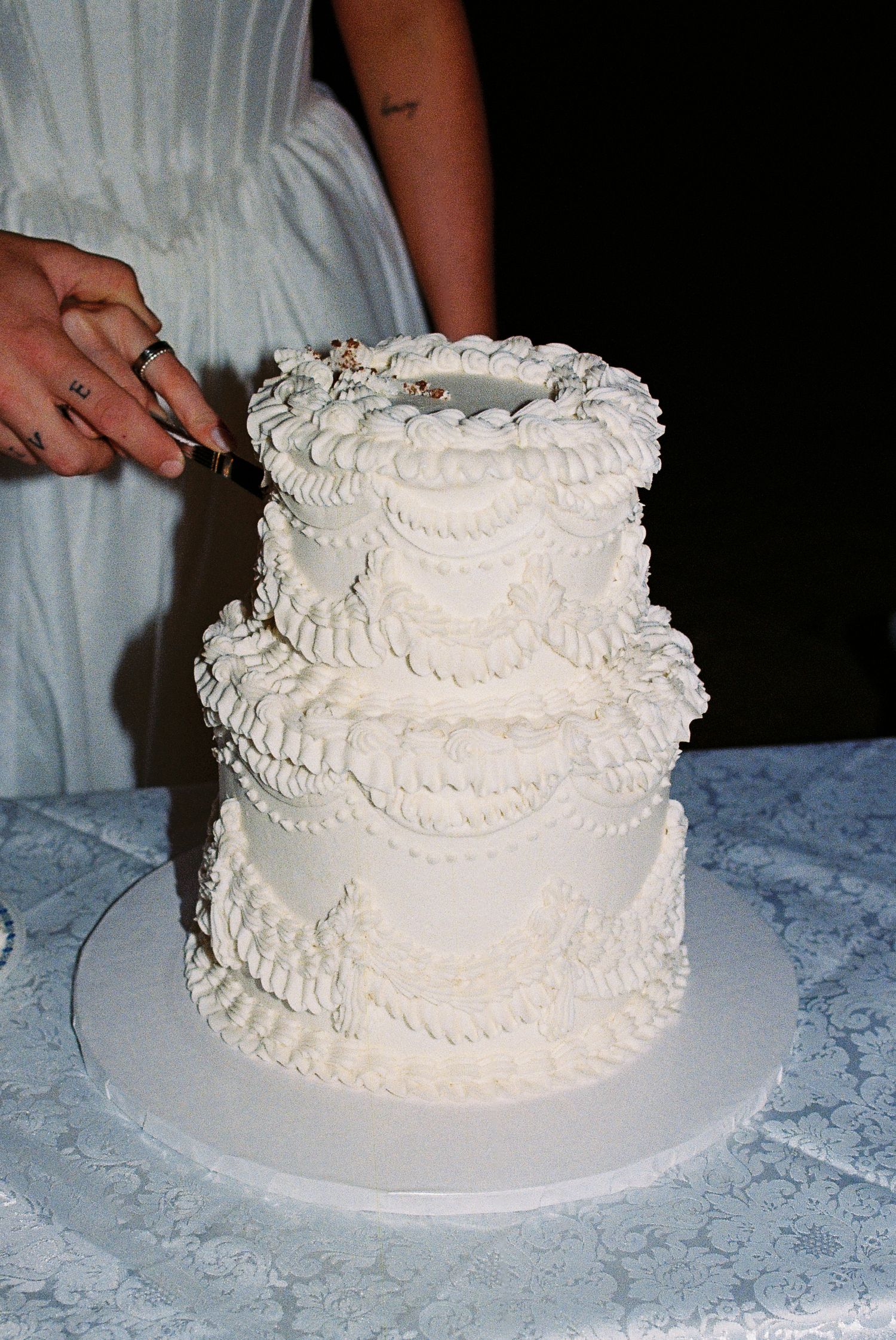 White two-tiered wedding cake with intricate icing designs around the whole cake. The bride and groom are cutting the first slice.