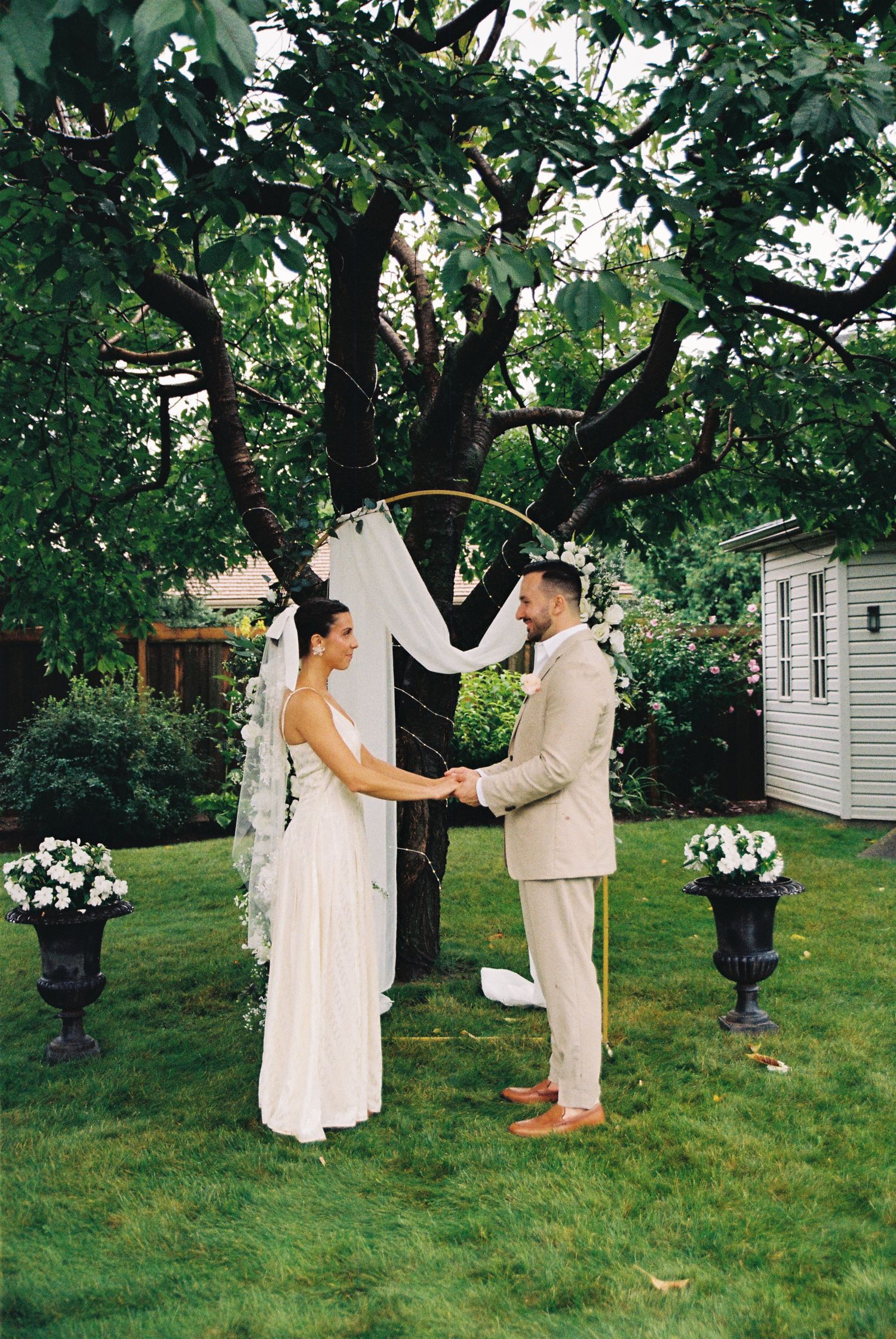 Bride and groom holding hands in front of altar during a backyard ceremony captured on 35mm film. Vivid green grass a tree surrounds them.