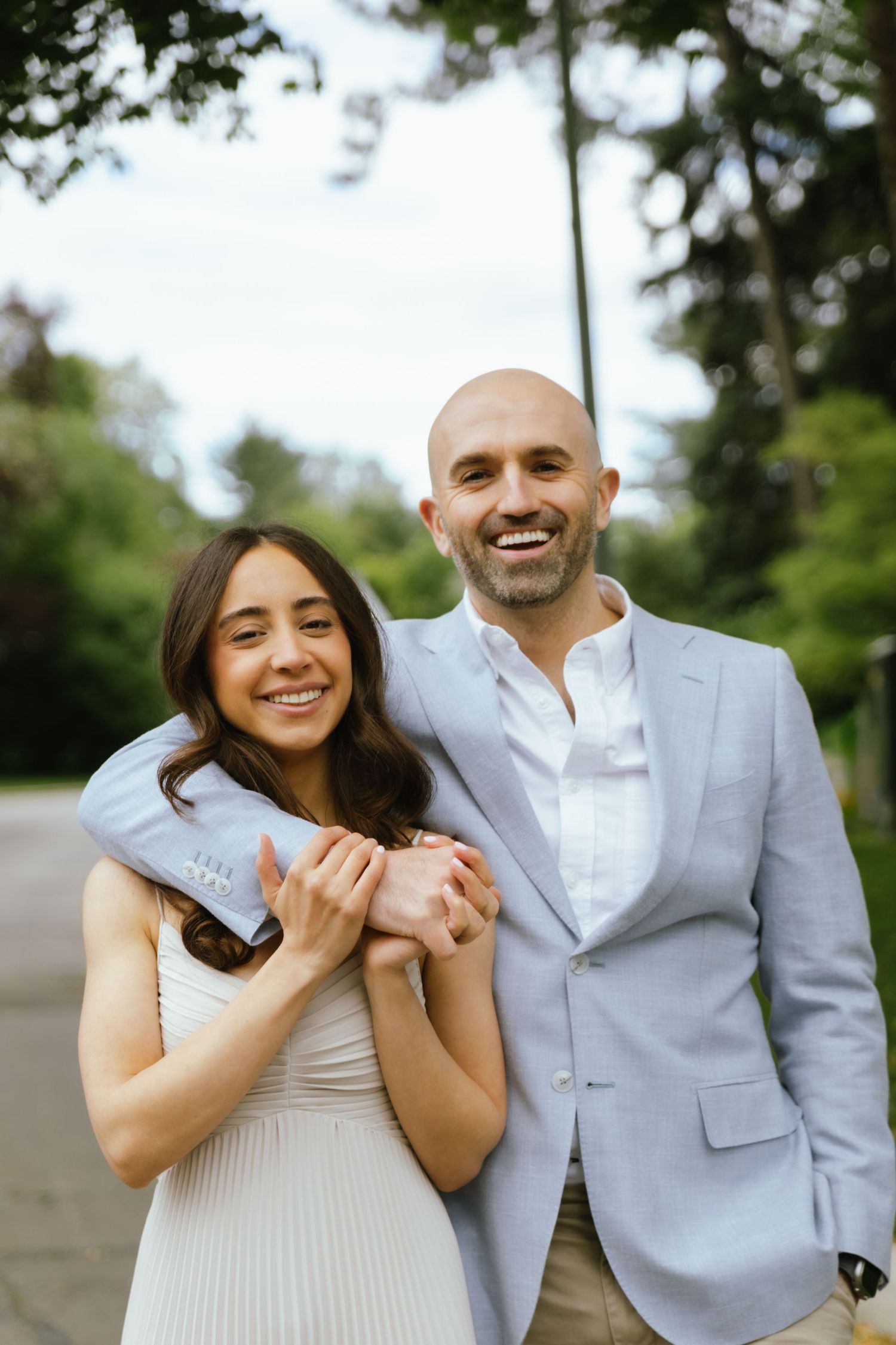Two fiancés pose together on a quiet street lined with greenery before their engagement party. They are both smiling.