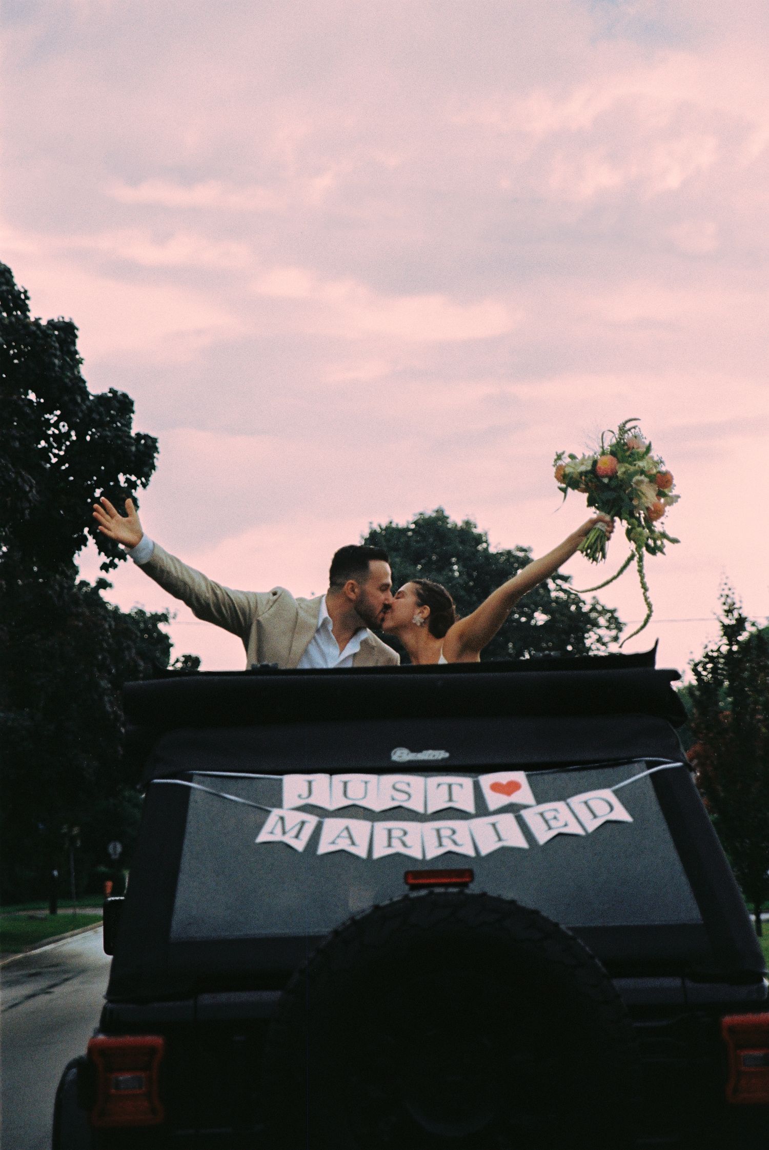 Bride and groom pop out the sunroof during sunset to kiss. The bride holds her bouquet and the back of the car says 