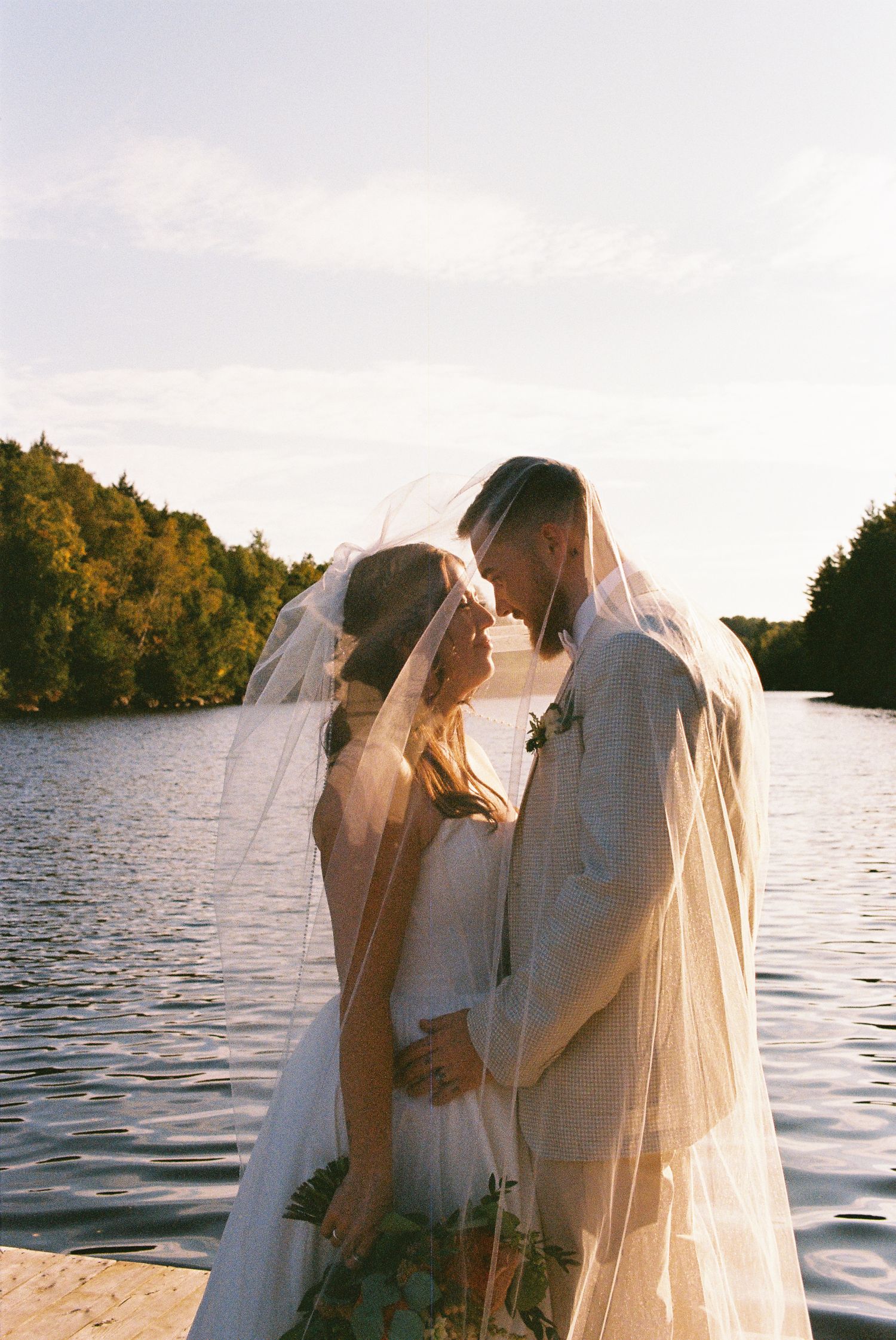 Bride and groom underneath the bride's veil standing on a dock in front of a lake during golden hour. Captured on 35mm film.