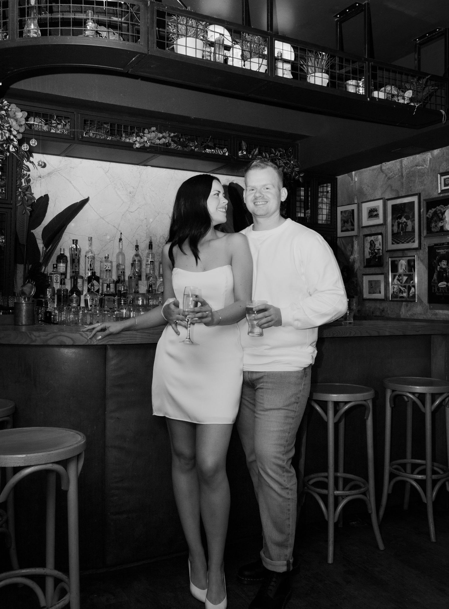 Fiancés posed together a bar where they met in Toronto, Ontario. The woman stares lovingly at her partner and the man smiles at the camera.