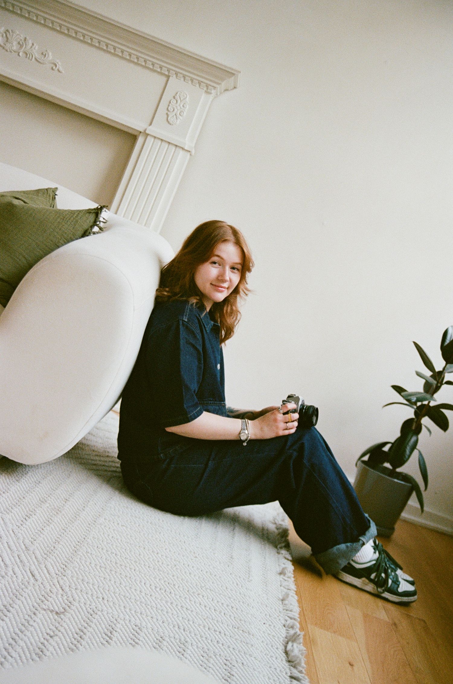Photographer holding a film camera sitting on a carpet next to a couch.