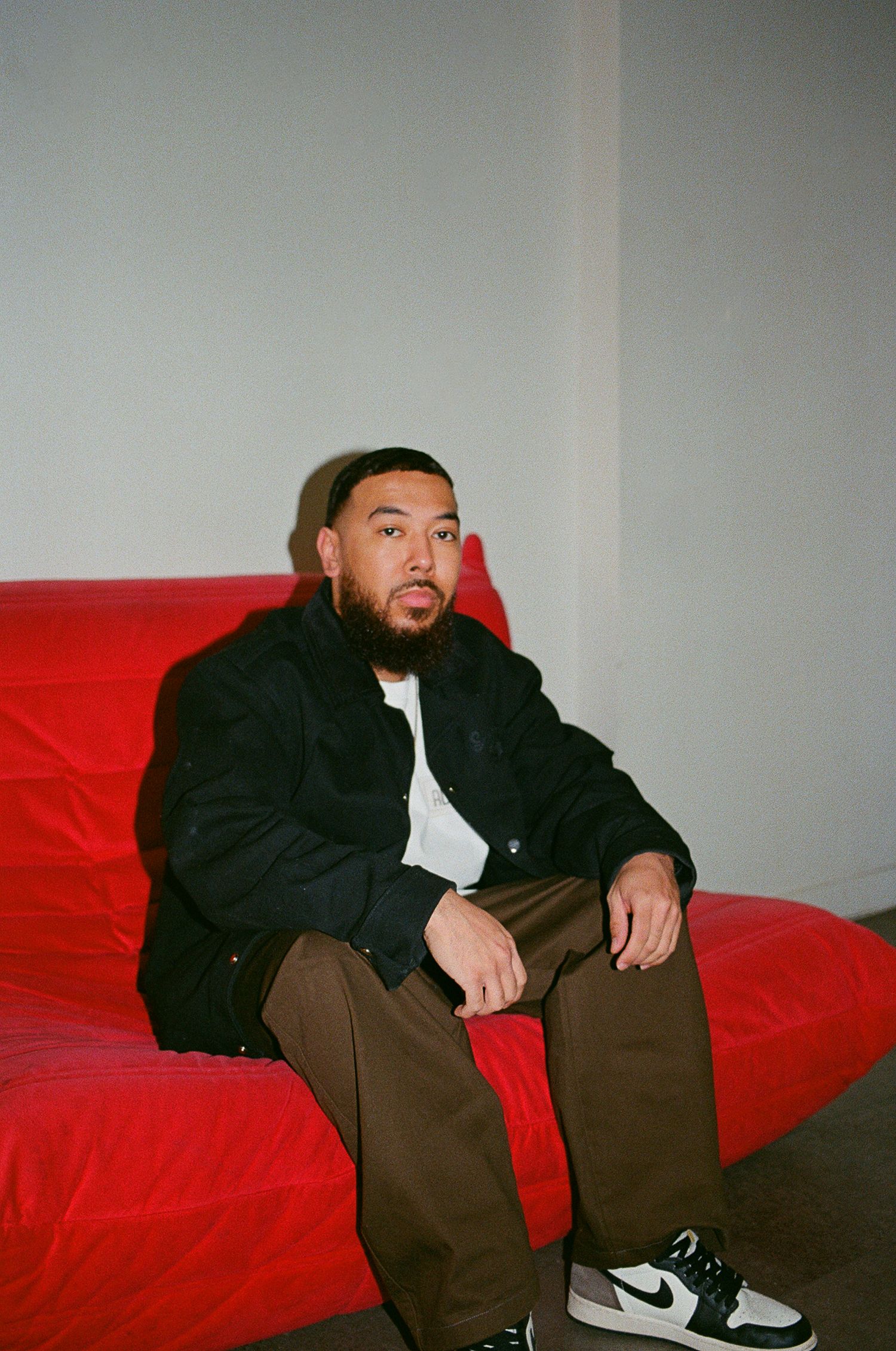 Film portrait of man sitting on a red couch wearing a black jacket and brown pants.