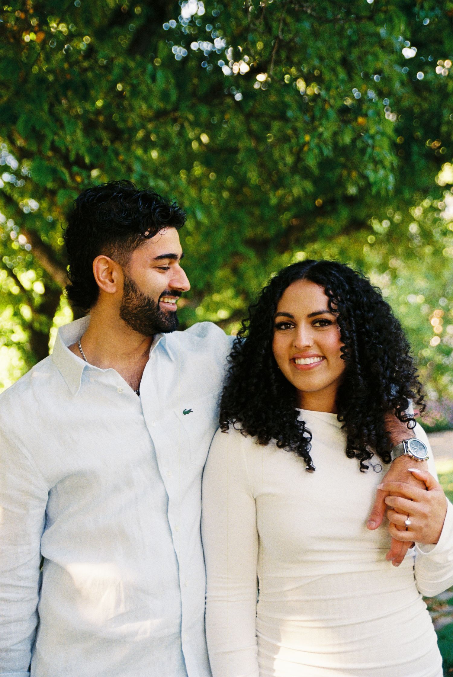 Fiancés in the Toronto Music Gardens dressed in white. They are both smiling and the man is staring lovingly at his partner.
