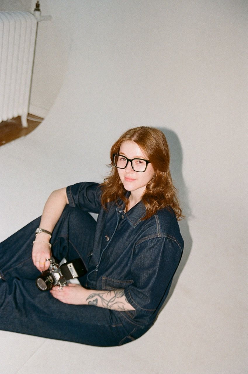 Photographer Shayla Anderson posed in the studio with a film camera in her hand and wearing a matching denim pant and button up shirt set.