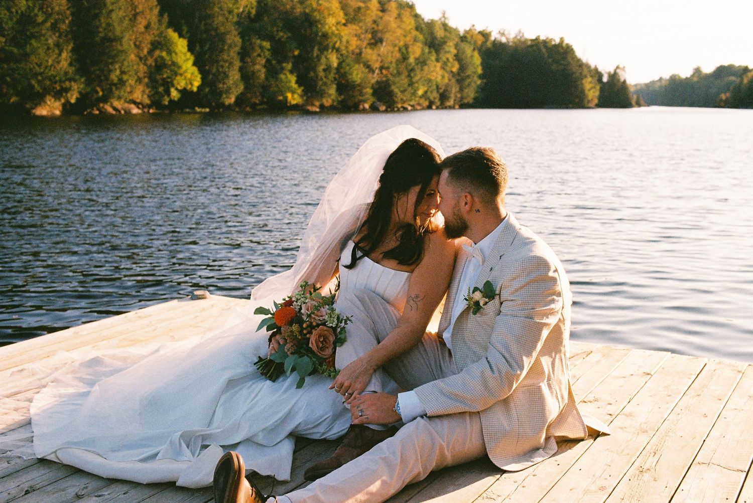 Bride and groom sitting together on a dock next to a lake with their foreheads touching. Captured on 35mm film.
