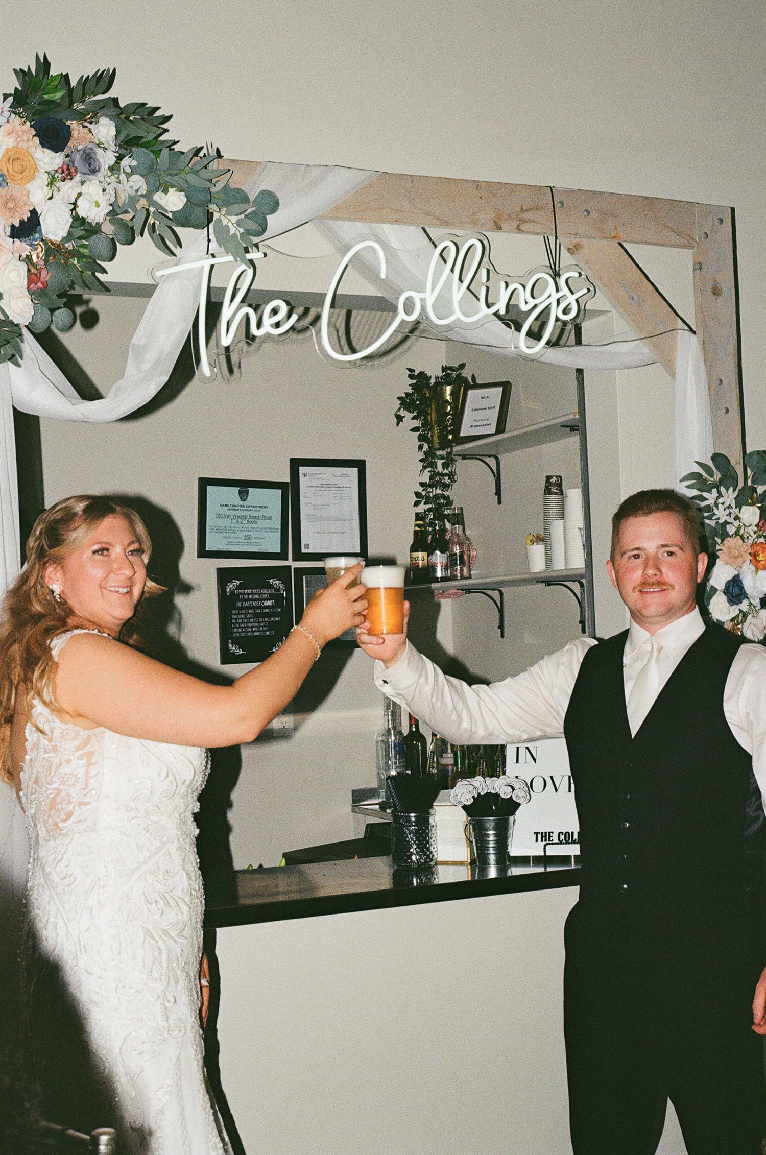 The bride and groom at a wedding are cheers-ing drinks next to the bar with a light up sign displaying their last name that hangs overhead.