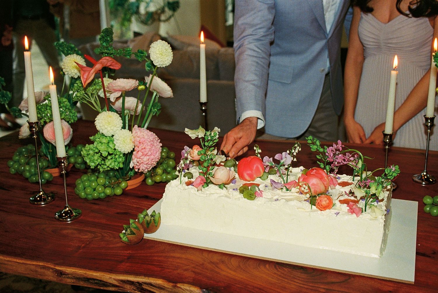 Fiancés cut their cake together at their engagement party. The cake is adorned with green and pink florals and surrounded by tapered candles
