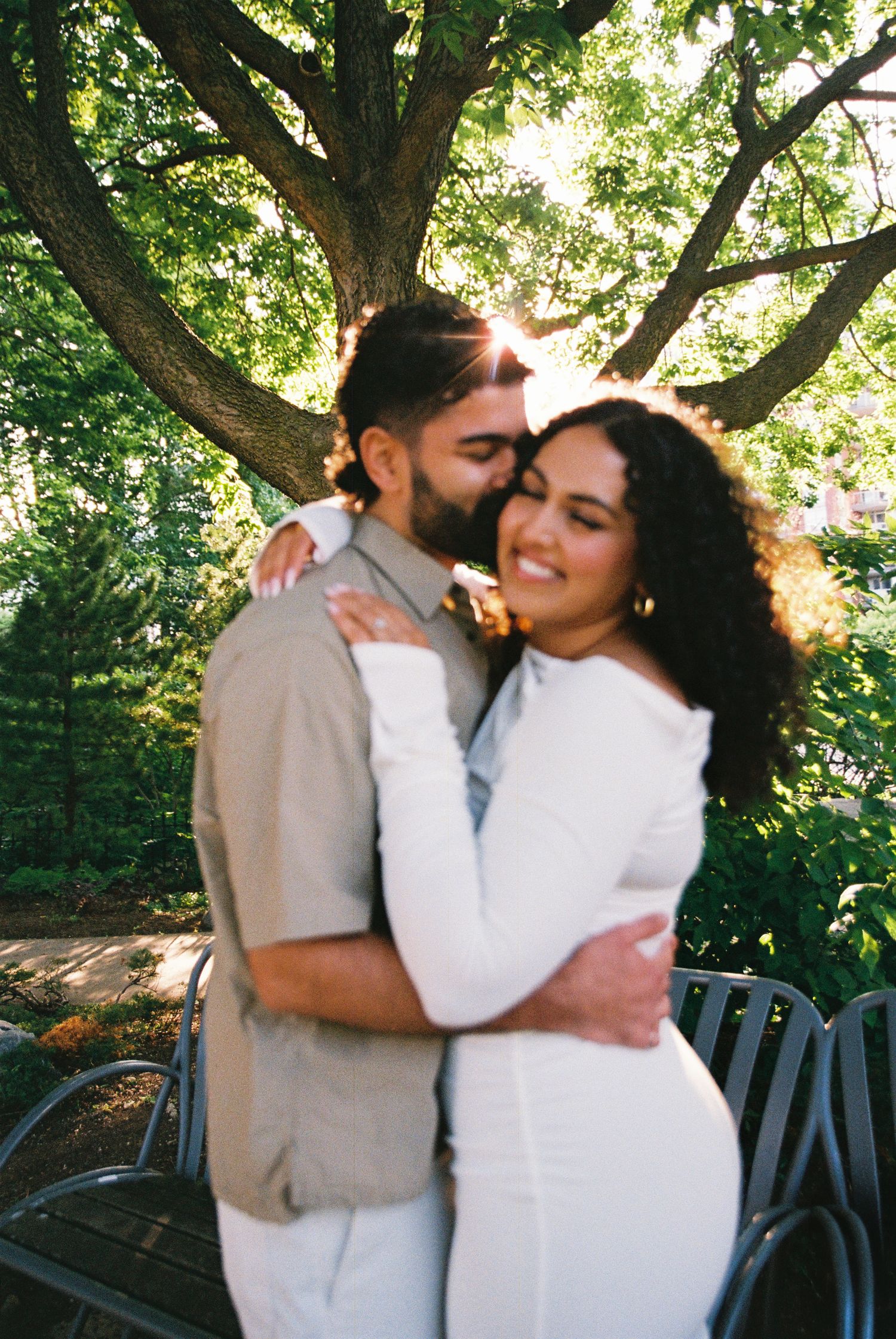 Fiancés standing in the Toronto Music Gardens together for their engagement photos. The man kisses the woman on her cheek.