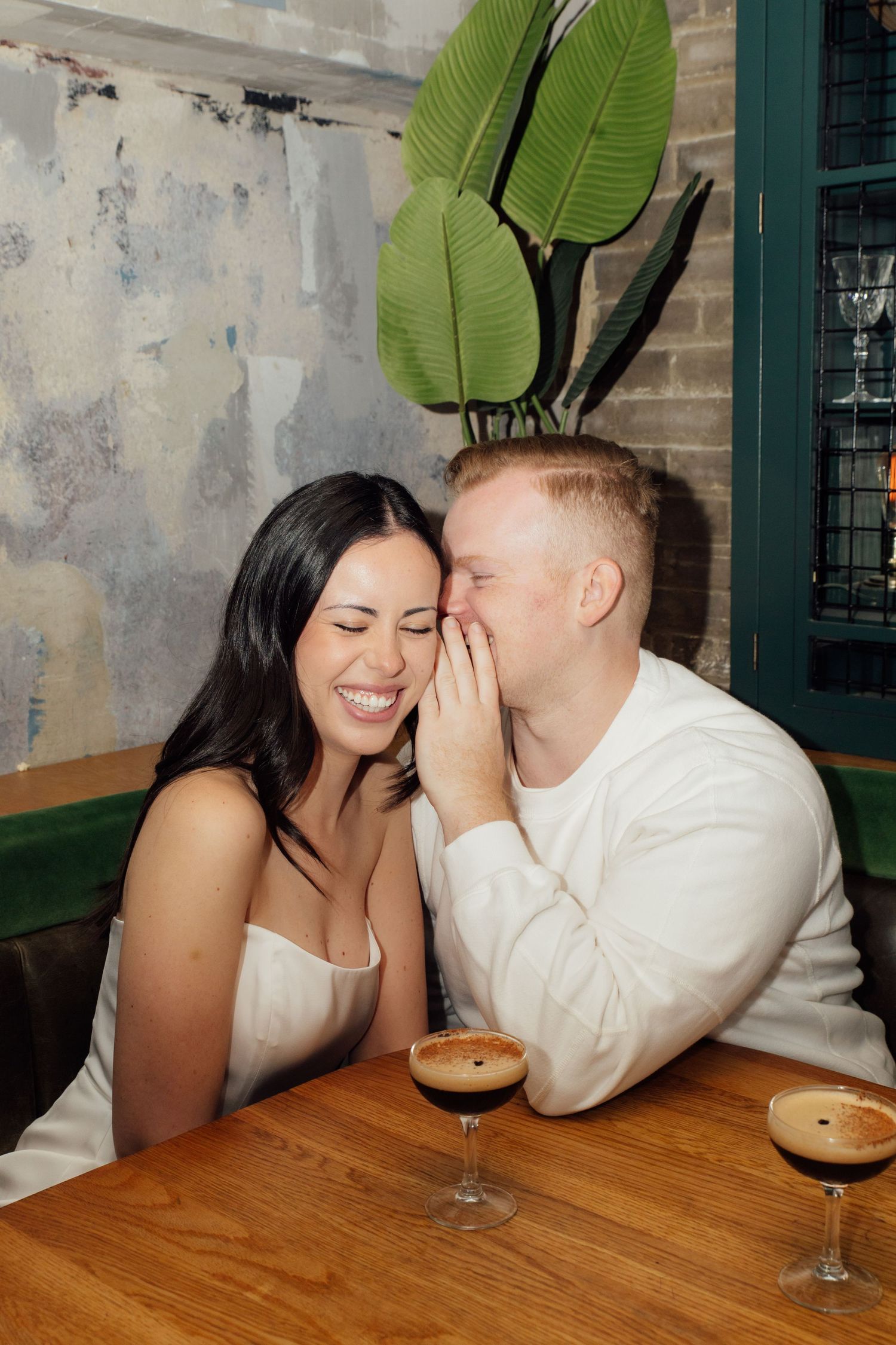 A couple sits together at a booth with espresso martinis, the man is covering his mouth and telling the woman a secret while she smiles.