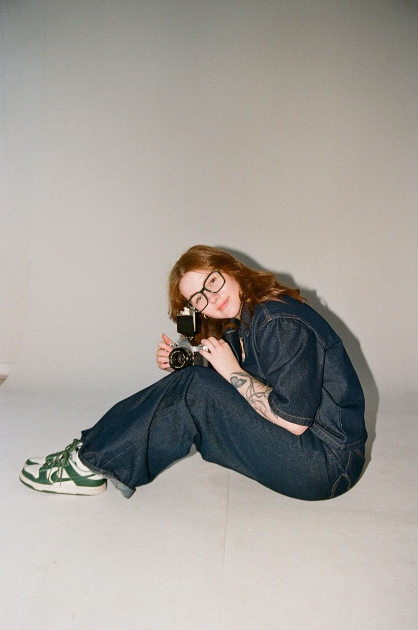 Photographer Shayla Anderson posed in the studio with a film camera in her hand and wearing a matching denim pant and button up shirt set.