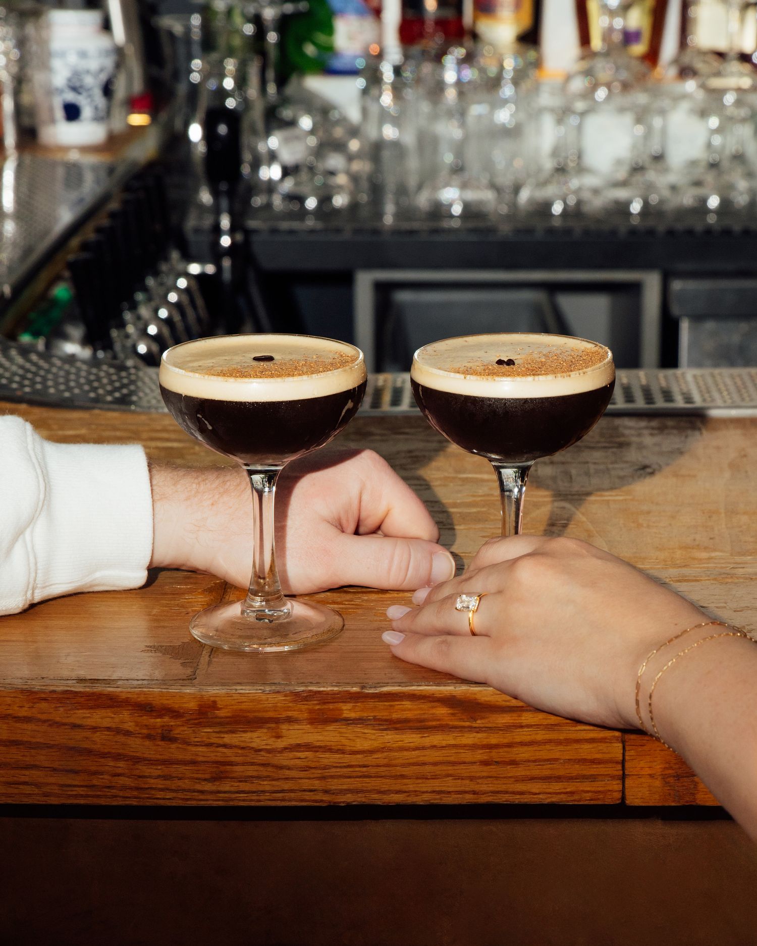 Two hands next to two espresso martinis on top of a bar. The woman is wearing an engagement ring.