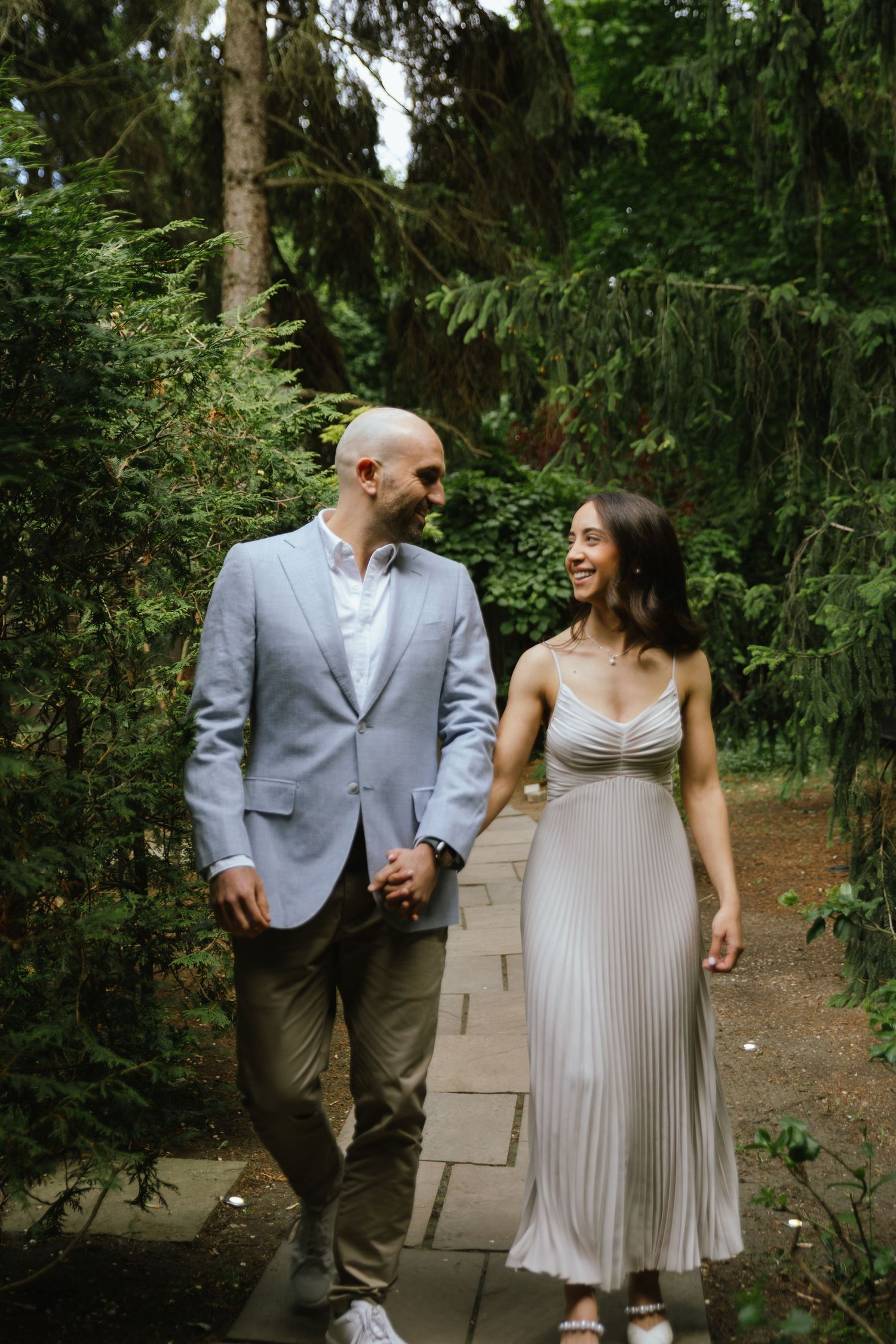 Couples portrait of fiancés holding hands, smiling, and looking into each others eyes as they walk towards the camera in a garden.