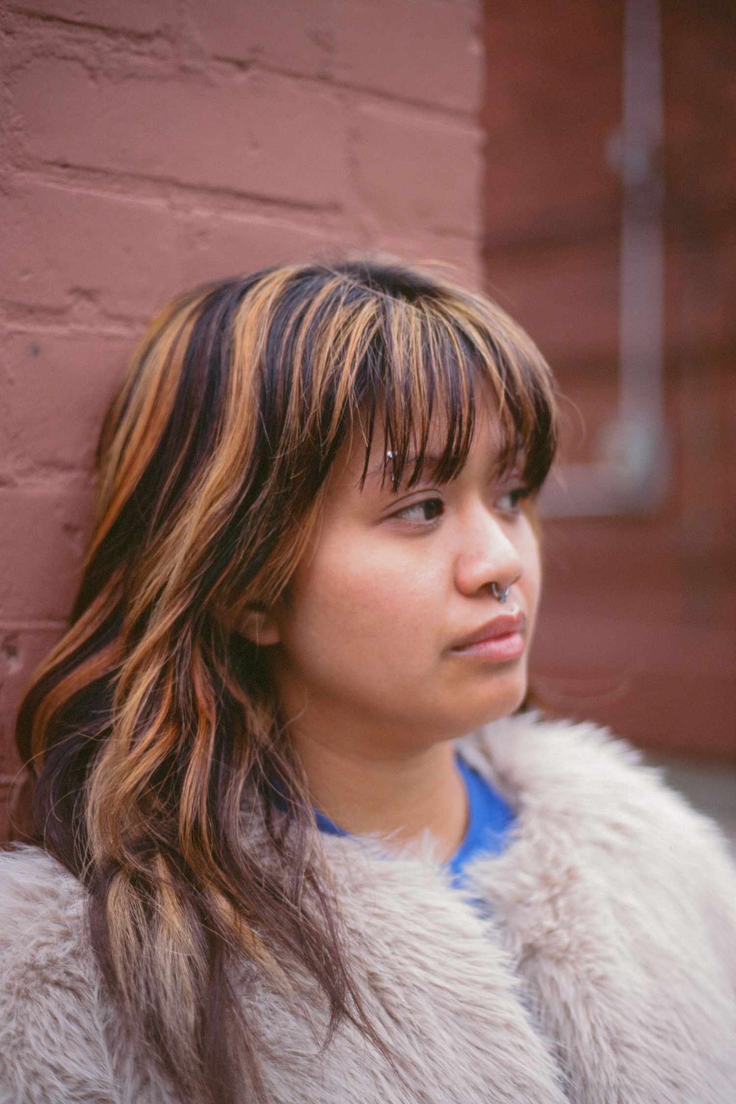 Film portrait of woman in front of red-orange brick wall. She wears a fur jacket, blue top, and is looking off into the distance solemnly.