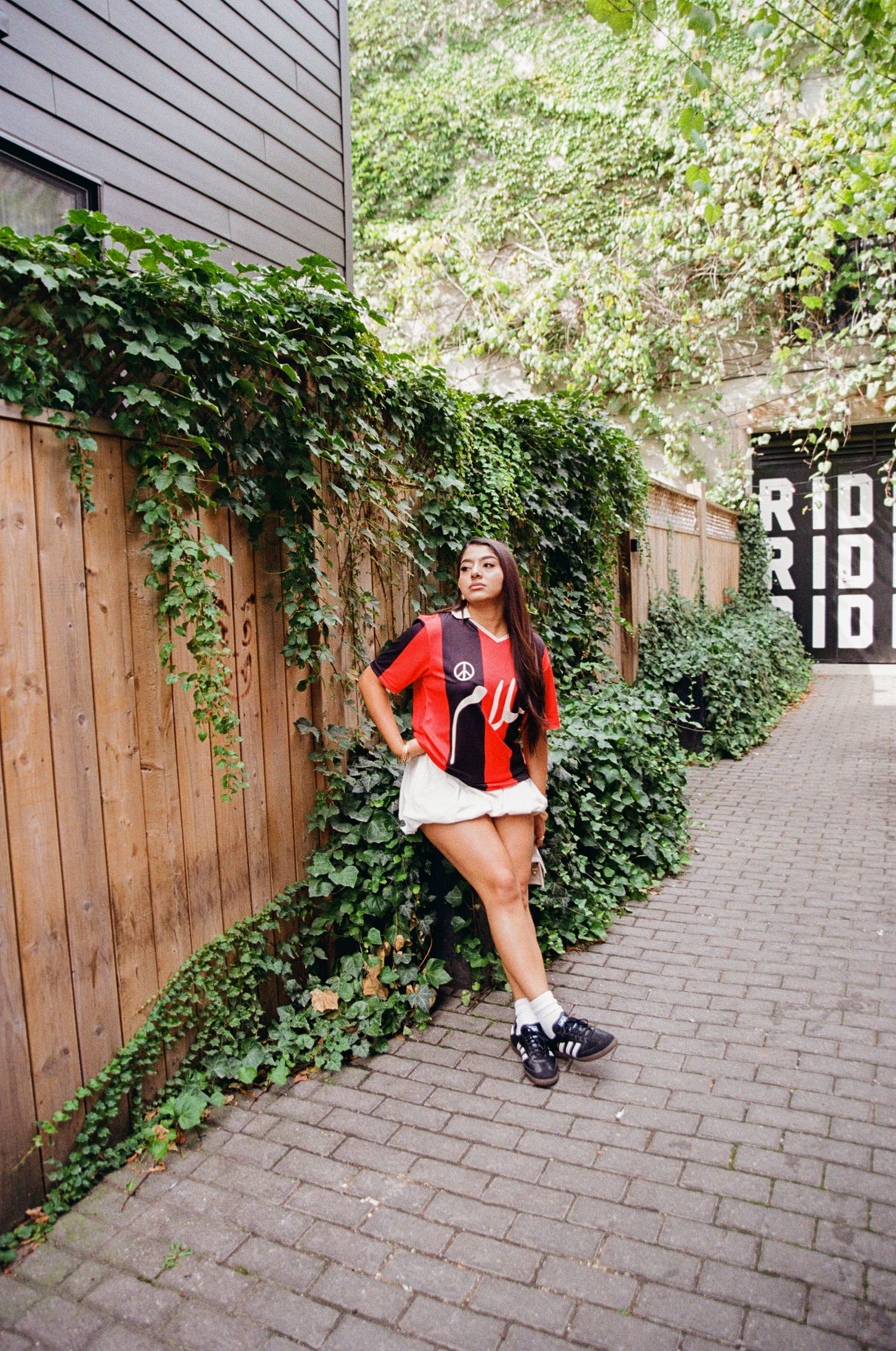 Film portrait of woman posed in front of a wooden fence and ivy, wearing a red and black striped jersey, sneakers, and a white puffy skirt.