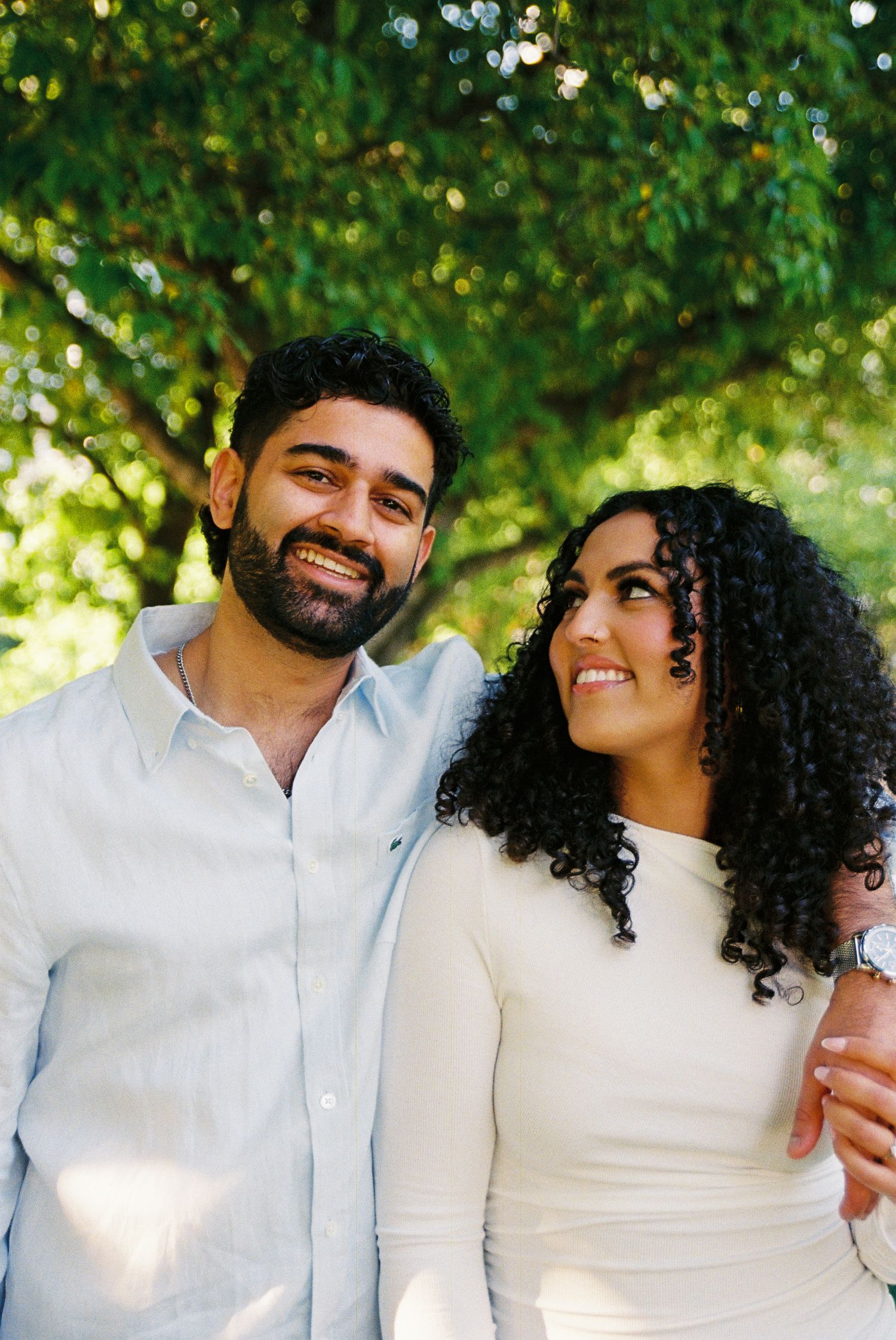Fiancés in the Toronto Music Gardens dressed in white and light blue. They are both smiling and the woman is starin lovingly at her partner.