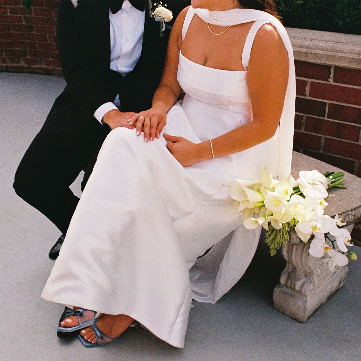 Groom wearing a black tuxedo and bowtie and bride wearing a satin long wedding gown and matching scarf. They are seated next to a bouquet.