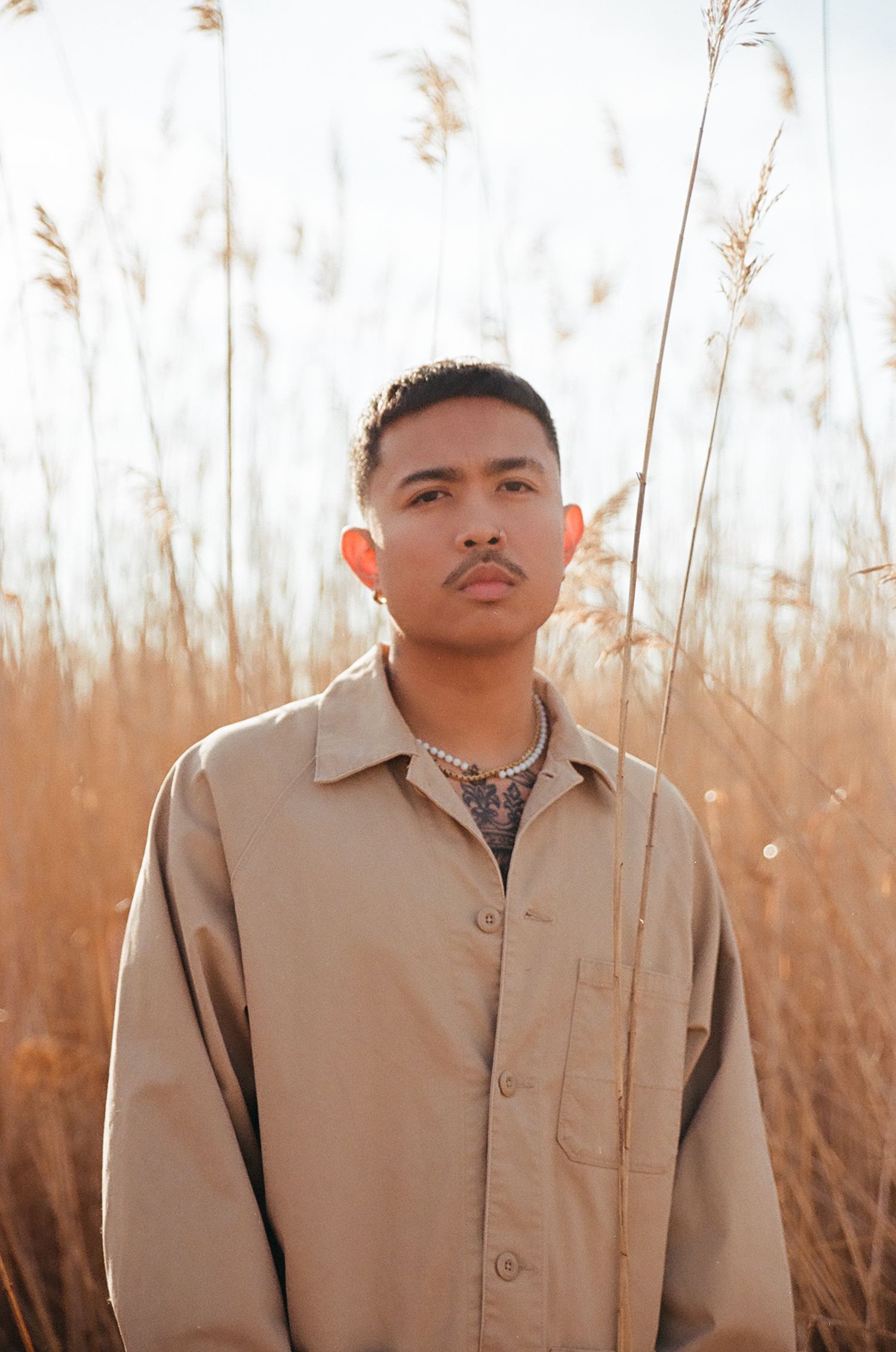 A man wearing a beige long sleeve button up shirt is standing in a wheat field during sunrise.