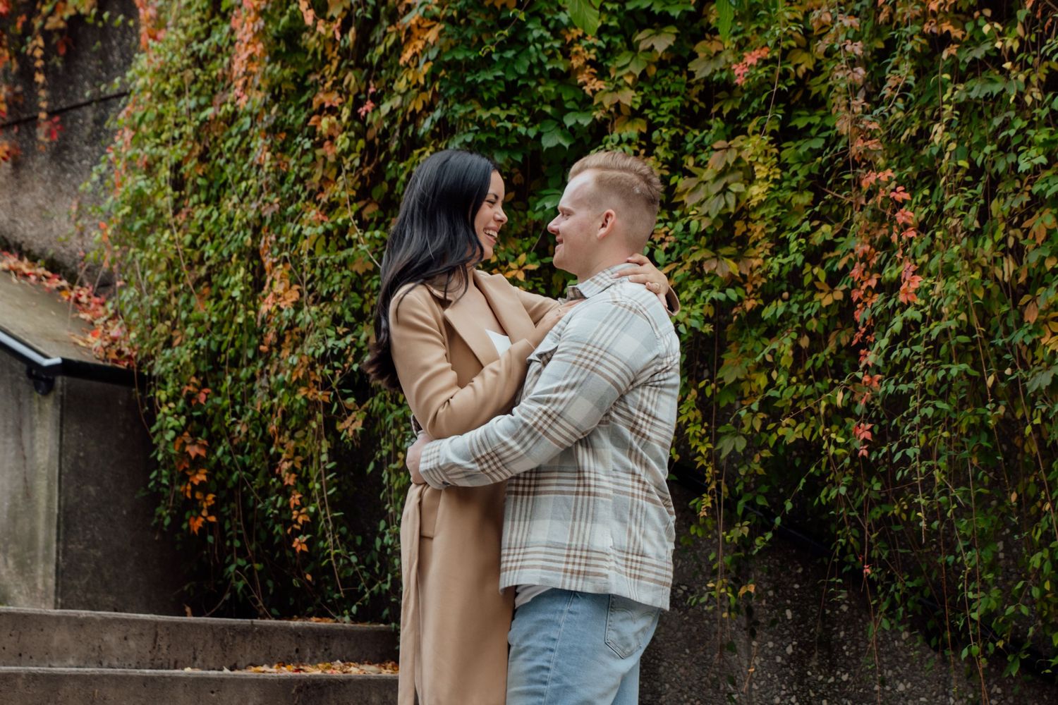 Fiancés hold each other in their arms as they stand outdoors on a set of stone steps in front of greenery, orange leaves, and ivy.