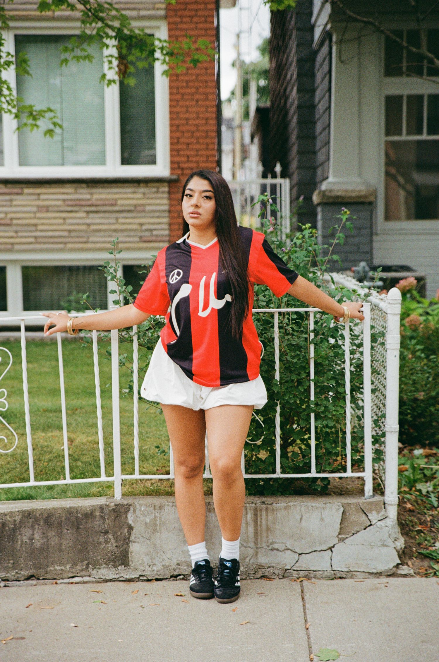 Film portrait of woman posed in front of white fence wearing a red and black striped jersey, sneakers, and a white puffy skirt.