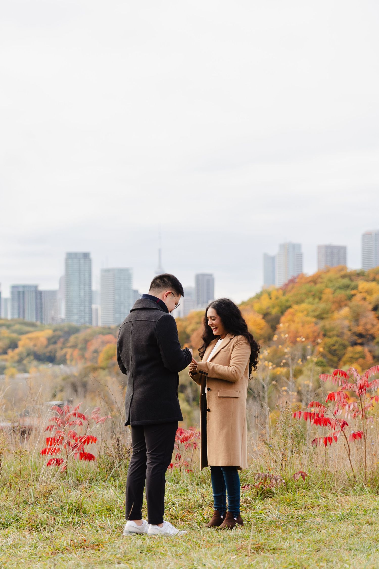A man proposes to his girlfriend atop a hill at Evergreen Brickworks surrounded by fall leaves. The CN tower and Toronto skyline are behind.