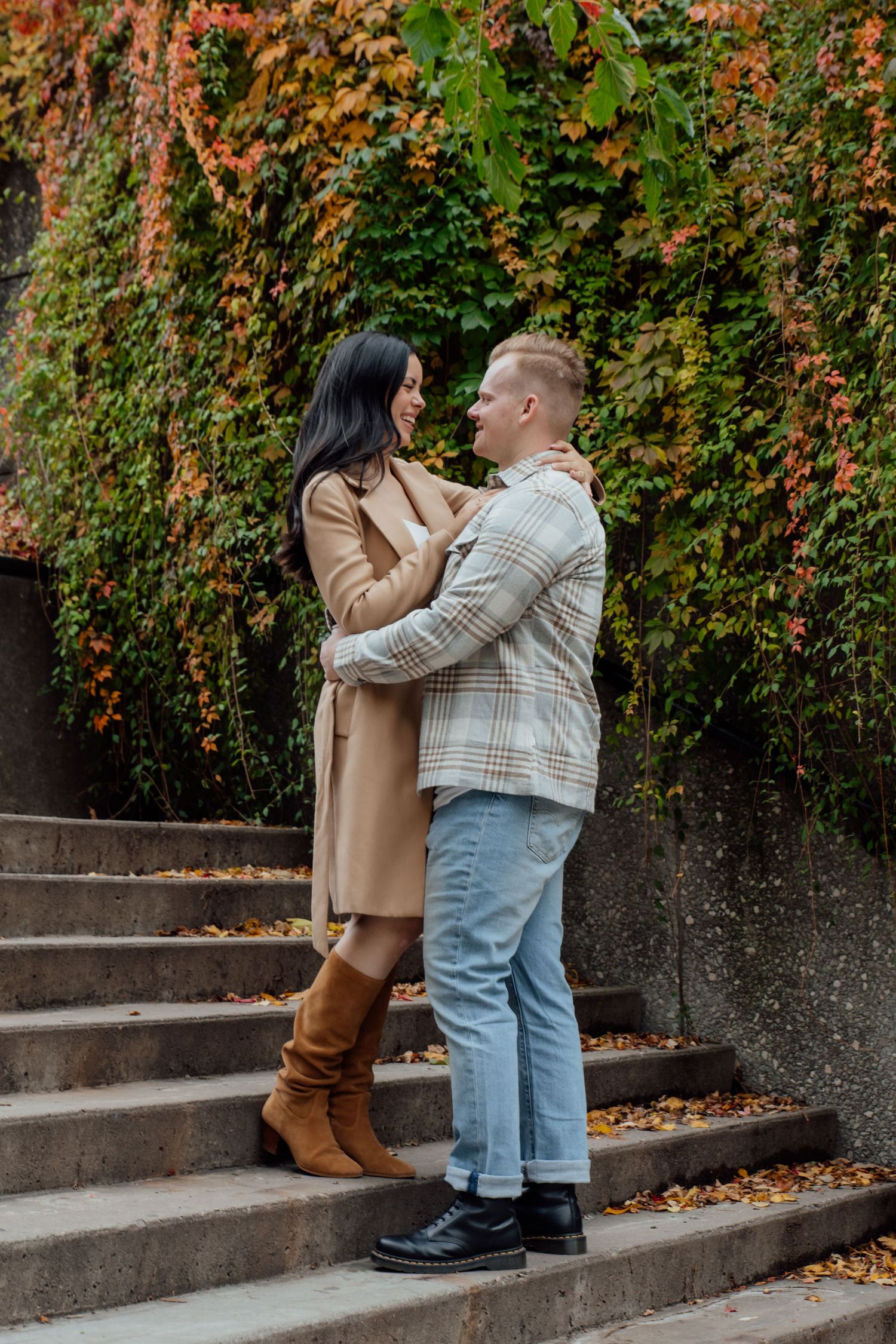 Fiancés hold each other in their arms as they stand outdoors on a set of stone steps in front of greenery, orange leaves, and ivy.