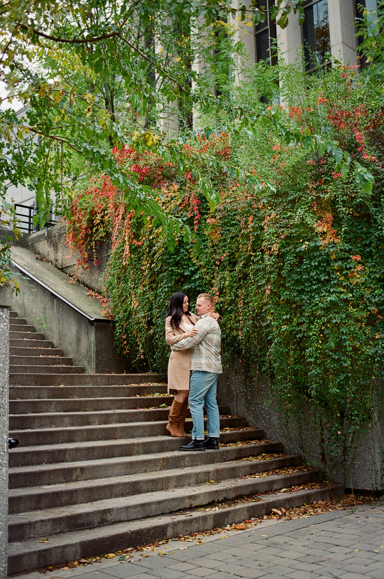 Fiancés hold each other in their arms as they stand outdoors on a set of stone steps in front of greenery, orange leaves, and ivy.